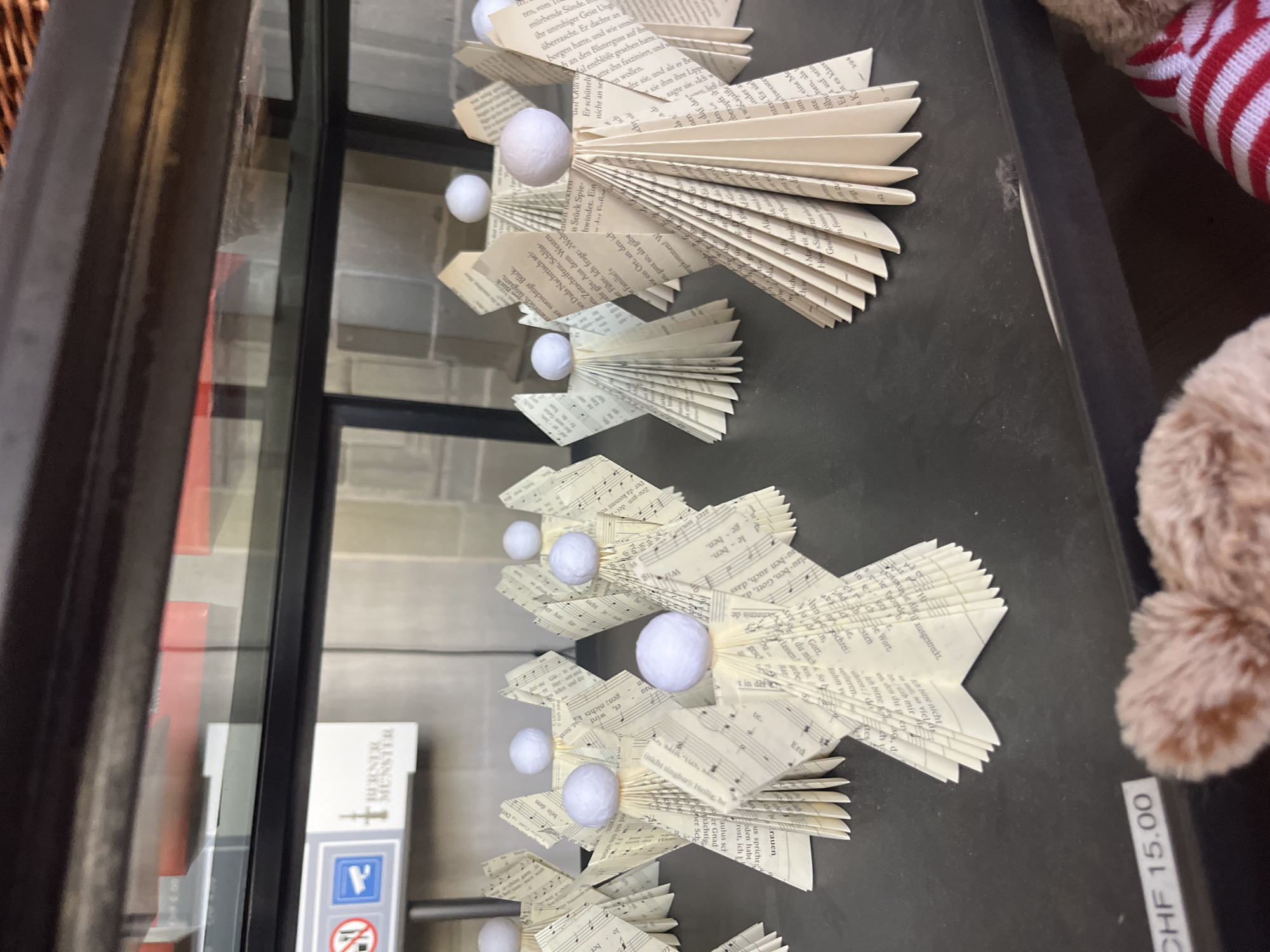 In Bern, Switzerland, a display case showcases a collection of handmade paper angels crafted from sheet music. The angels, arranged in rows, have bodies made of folded paper and spherical white heads. Some angels are positioned closer to the viewer, while others recede into the background. In the upper left corner of the display, a sign with a prohibition symbol and a surveillance camera icon is visible, accompanied by text that appears to read "BERNER MUNSTER". In the foreground, a price tag reads "CHF 15.00" next to a blurred image of a stuffed animal. The lighting suggests an indoor setting, possibly a shop or museum.