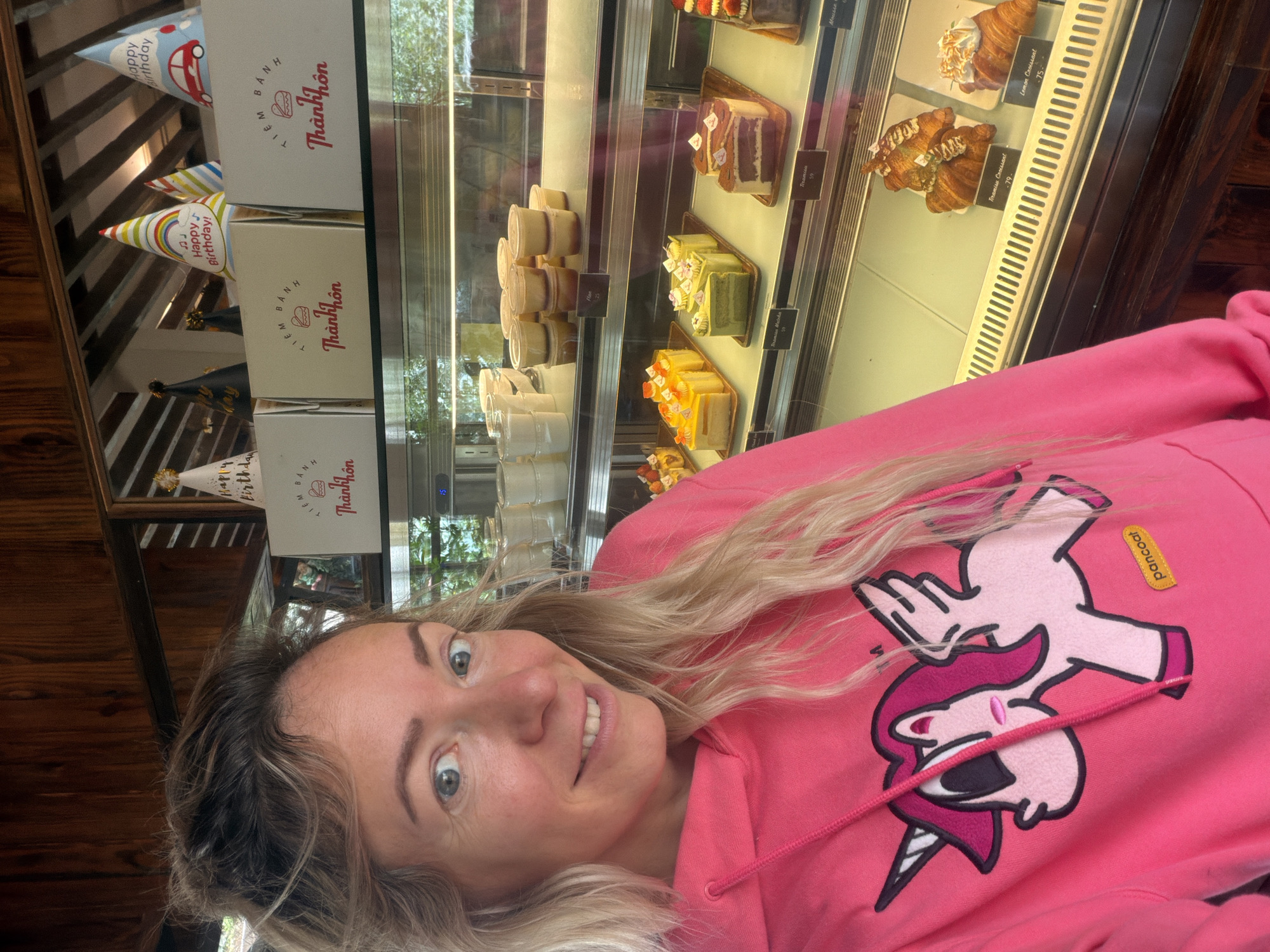 A woman with blonde hair pulled back into a messy bun and loose strands framing her face poses for a selfie in front of a bakery display case. She is wearing a bright pink hoodie with a graphic of a pink unicorn with wings on the front. Her eyes are wide and blue, and she has a slight smile. The display case contains an assortment of pastries, including cakes and croissants. Behind the woman, on a shelf above the display case, are several white boxes with Vietnamese writing and festive birthday hats. The background appears to be a wooden interior, suggesting the location is a bakery in Da Lat, Vietnam, known for its European-style architecture and numerous cake shops.