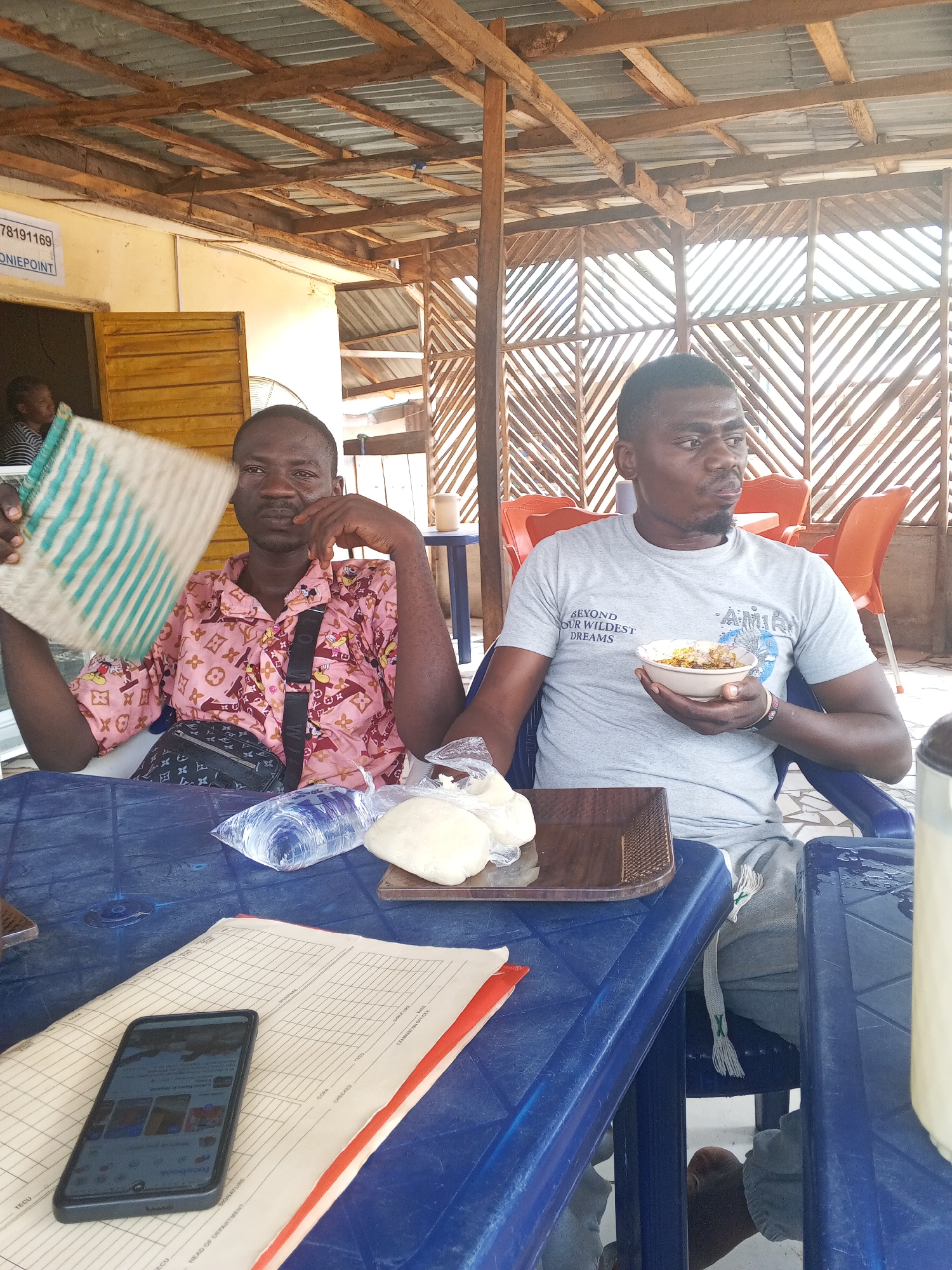 In Lokoja, Nigeria, two men are seated at a blue plastic table under a wooden and corrugated metal roof. The man on the left wears a pink patterned shirt and holds a woven mat. The man on the right wears a grey t-shirt and holds a bowl of food. On the table are a plastic bag of water, a tray with what appears to be dough, a stack of papers, and a smartphone displaying a social media app. The environment suggests an outdoor eatery.