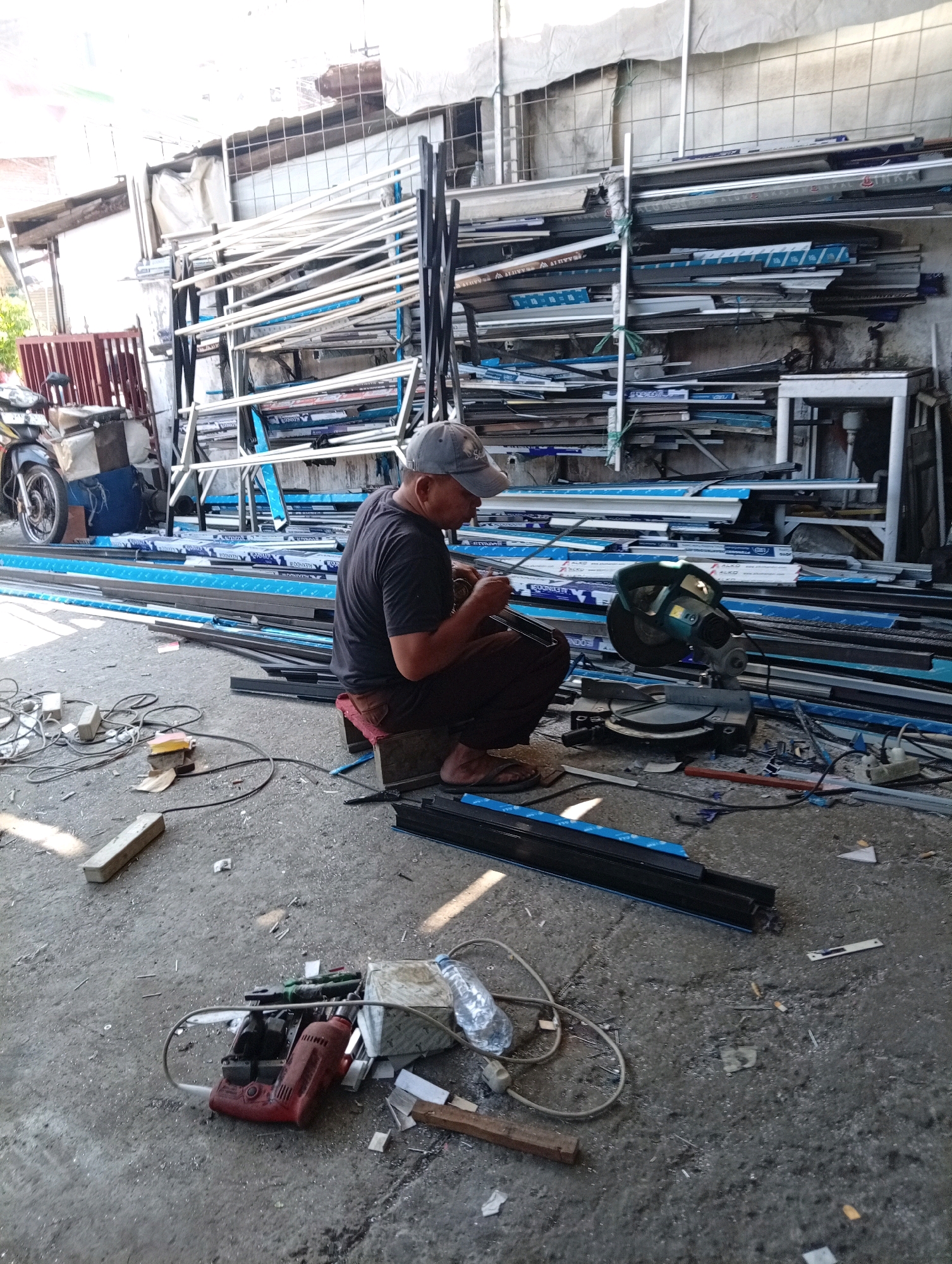 A man in a grey baseball cap and a dark t-shirt is sitting on a small red stool in an outdoor workshop in Bangah, Indonesia. He is focused on a piece of metal he is holding with his hands, appearing to be working on it. In front of him, on the concrete ground, is a green and grey compound miter saw, along with various pieces of aluminum framing and tools, including a red power drill and a plastic water bottle. The workshop is filled with stacks of aluminum profiles and other building materials. A motorcycle is parked on the left side of the image. The ground is littered with debris, including metal shavings and discarded materials. The overall environment suggests an active, albeit somewhat cluttered, workspace.