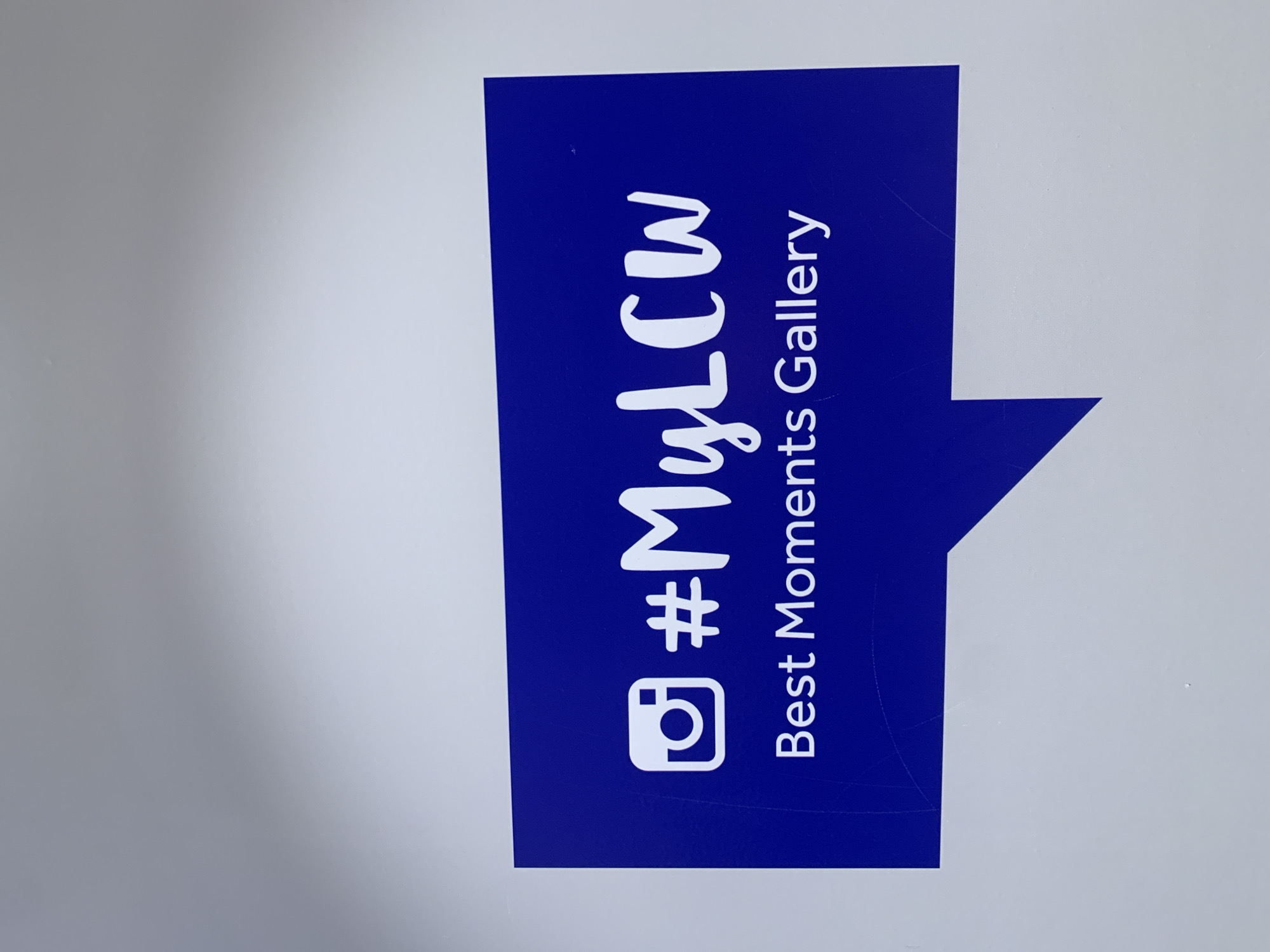 The image shows a blue sign with white text. The text includes an Instagram icon, the hashtag "#MyLCW", and the words "Best Moments Gallery." The sign is positioned on a white surface. The image provides no information to determine the location of the sign.