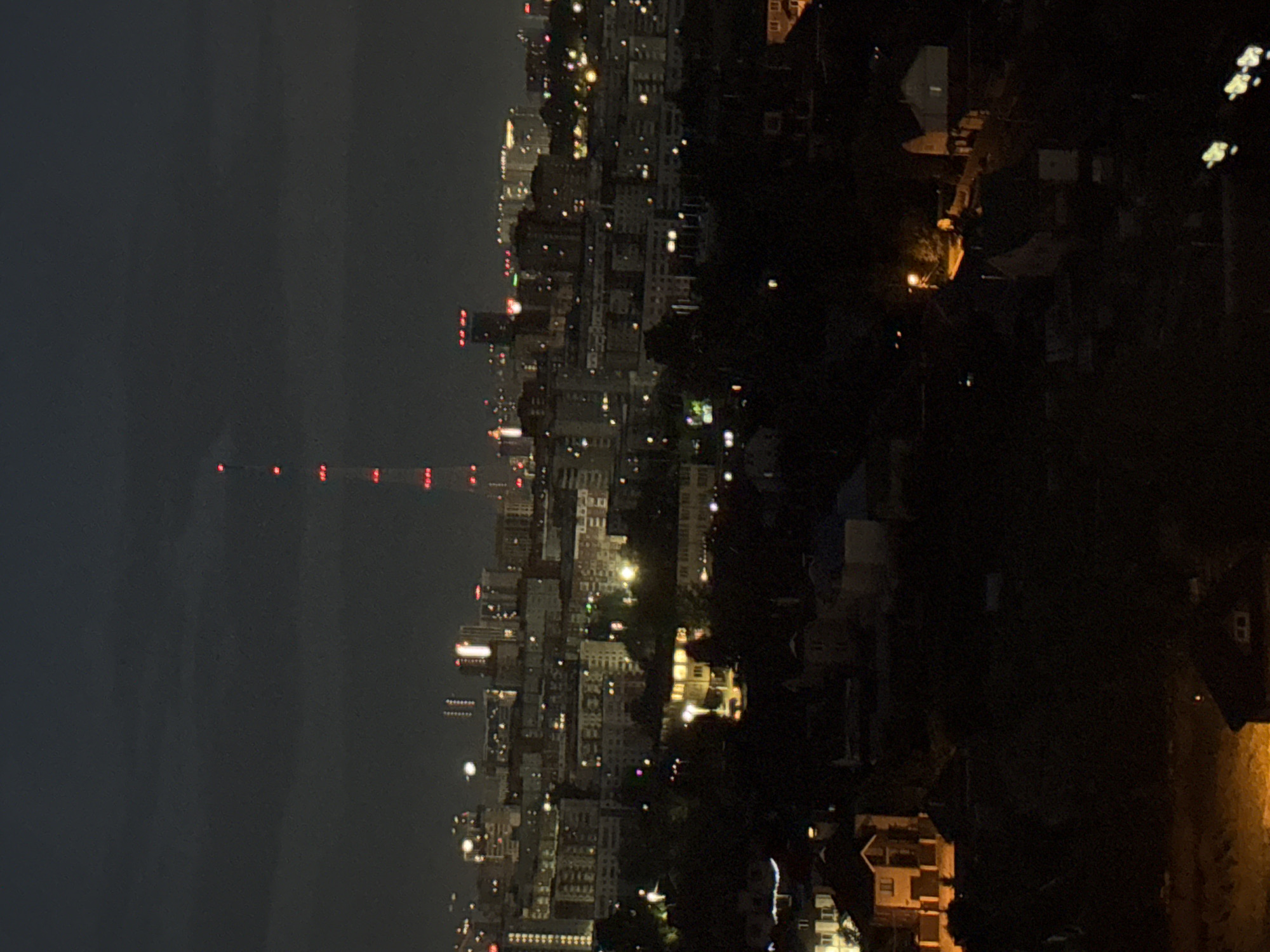 This is a nighttime aerial view of a city skyline, likely Perm, Russia, characterized by a dark, overcast sky. A prominent telecommunications tower, illuminated with red lights at intervals, stands tall in the background. The middle ground is filled with a dense urban landscape of apartment buildings and other structures, many displaying illuminated windows. In the foreground, the scene transitions to a more sparsely populated area with houses, some with lit windows and streetlights casting warm glows. The overall impression is a sprawling urban environment at night, with the telecommunications tower serving as a central landmark.