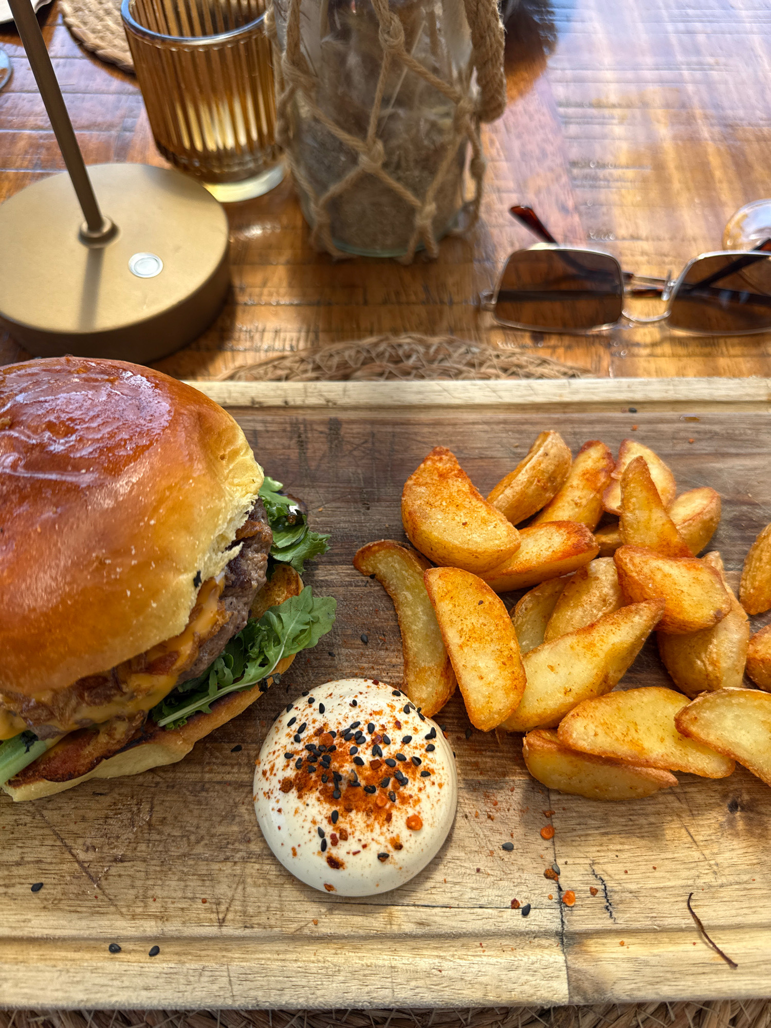 An overhead shot shows a wooden table with a burger and fries, served on a wooden board. The burger has a golden-brown bun, a thick patty, and green lettuce. The fries are wedge-shaped and seasoned with paprika. A dollop of white sauce, topped with black seeds and red pepper flakes, sits beside the burger. In the background, a glass vase with rope detailing and sunglasses are visible on the table. The scene appears to be set in a restaurant in Vila Nova de Gaia, Portugal, possibly overlooking the Douro River or the city of Porto.