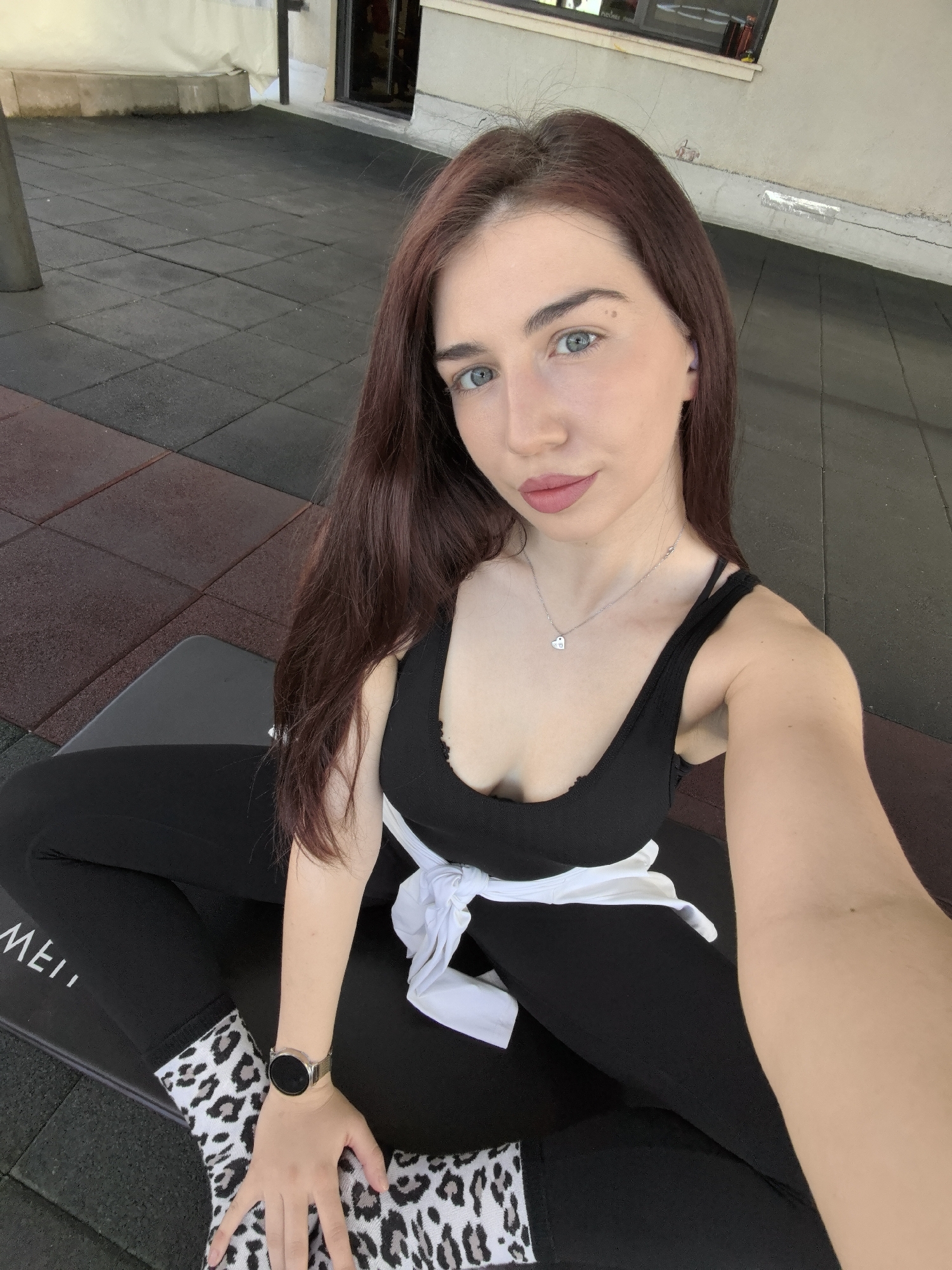 A daytime selfie captures a young woman seated in what appears to be an outdoor or semi-outdoor gym area in Stara Zagora, Bulgaria. She has long, dark brown hair and light blue eyes, looking directly at the camera with a neutral expression and slightly parted, pink-tinted lips.
She is dressed in a black sleeveless top or sports bra and black leggings. A white shirt is tied around her waist. Her socks are a notable detail, featuring a black, white, and grey leopard print pattern. Accessories include a silver-toned smartwatch with a black circular face and mesh band on her left wrist, and a delicate silver chain necklace with a small heart-shaped pendant.
The setting includes a floor covered with square rubber tiles in black and a reddish-brown color. In the background, a light-colored wall or building with a dark window or doorway is visible. Part of the text "MEI" can be seen printed on her left legging.