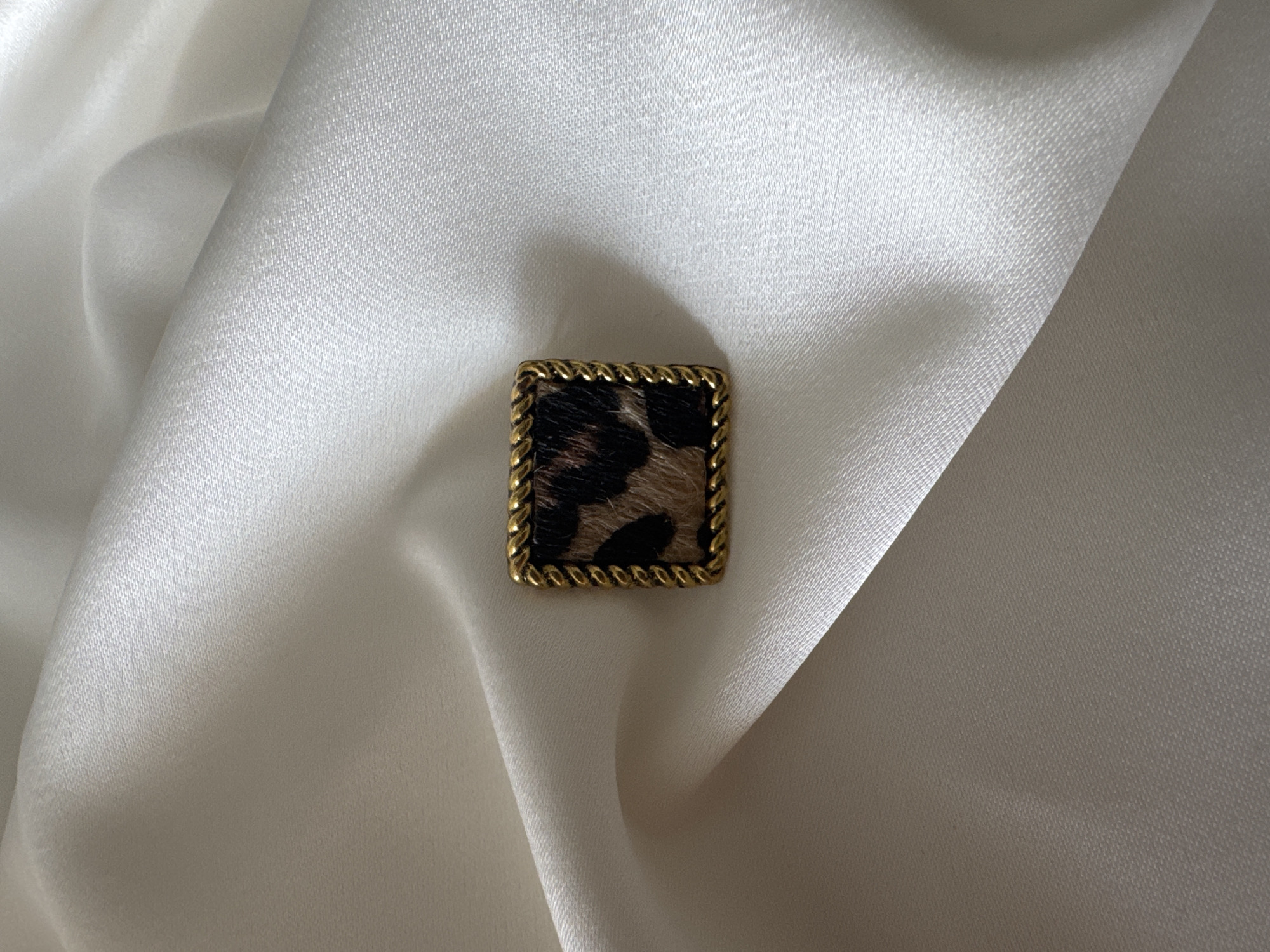 A square leopard-print brooch with a gold twisted rope border rests on a wrinkled white satin fabric. The brooch is centrally located on the fabric, which is draped and folded, creating soft shadows. The background is plain white silk fabric. The image is a close-up shot, focusing on the brooch. No people are visible in the image. The location is not specified.