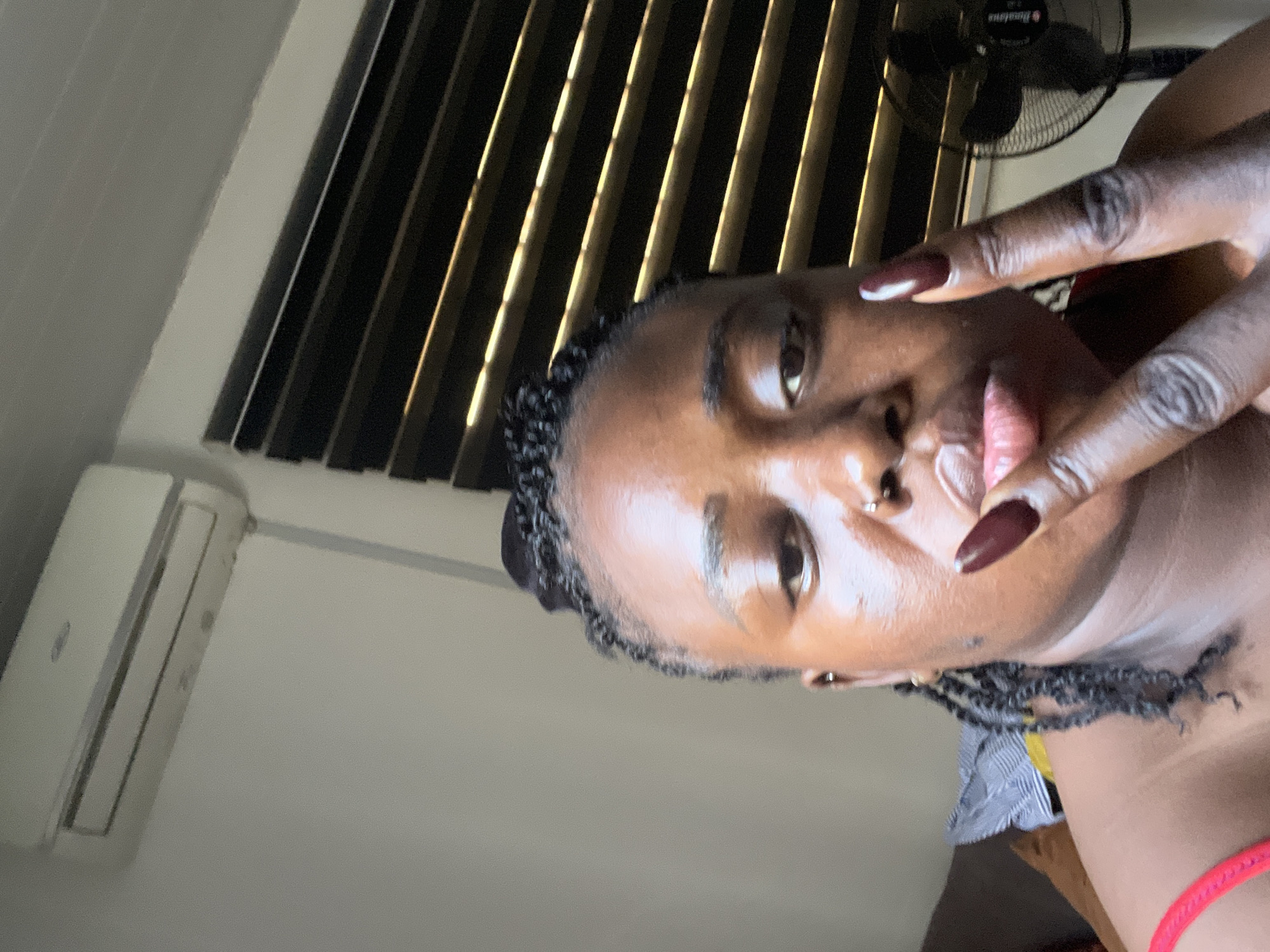 A close-up, selfie-style shot captures a Black woman with braided hair and long, dark red fingernails. She is positioned indoors, with her face filling most of the frame. Her head is slightly tilted, and her right hand is raised to her lips, with her index and middle fingers extended in a peace sign. She wears a red garment on her upper body. The background reveals a white wall with an air conditioning unit mounted near the ceiling. To the right, a dark-slatted window blind is visible. In the far right of the image, a black oscillating fan is partially in view. The lighting suggests an indoor setting, possibly during the day. The location is identified as Abuja, Nigeria.