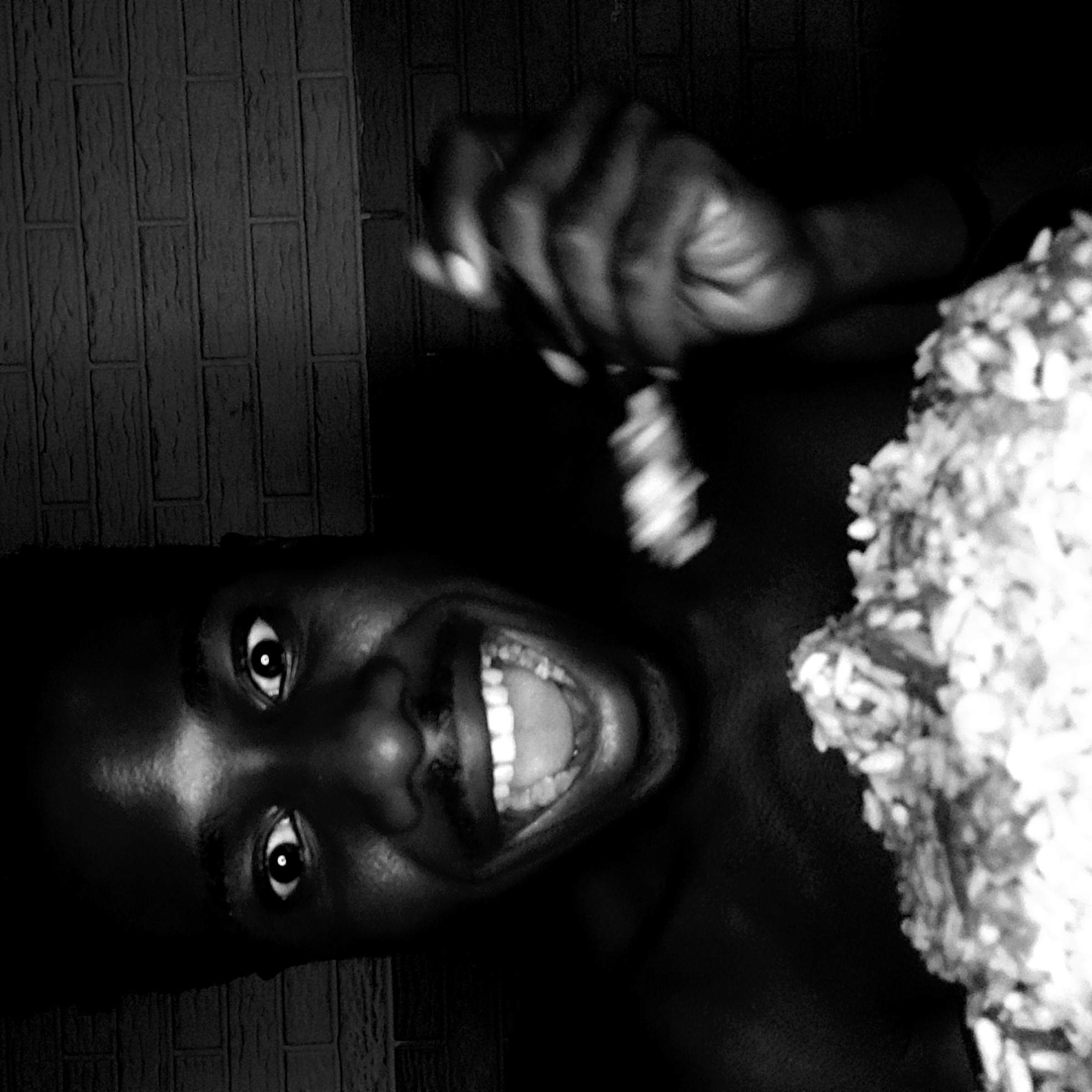 The black and white image features a person captured in a moment of extreme joy or excitement. The subject, whose face is tilted, gazes towards the viewer with wide, bright eyes and a dramatically open-mouthed smile, revealing a full set of teeth. Their expression is exuberant, almost ecstatic.
In the upper right portion of the frame, one of the person's hands is raised and appears blurred, suggesting motion, possibly holding a utensil or interacting with something just out of view. To the right of the person, a large, textured mound of food, resembling rice or grains, is prominently displayed, hinting at a meal being consumed or anticipated.
The overall scene is dimly lit, with a strong light source highlighting the person's face, creating deep shadows and high contrasts typical of a black and white photograph. The background is a dark wall featuring a subtle brick-like or patterned texture. There is no visible text. The image's intensity and the subject's expression convey a powerful sense of happiness or surprise, likely associated with the food. The location context for this media file is Narayi, Nigeria.