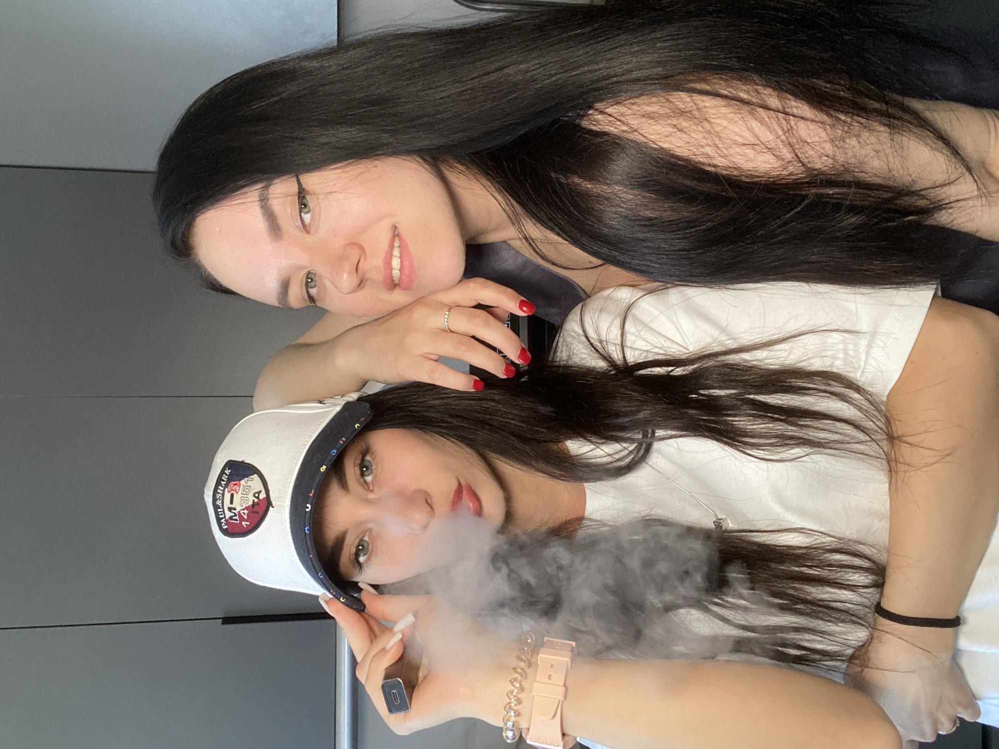 Two young women are posing for a photograph. One woman with long dark hair is lying on top of the other, smiling. The woman beneath is wearing a white t-shirt and a white baseball cap with a blue brim and an emblem on the front. She is exhaling vapor from a vape pen she is holding in her right hand. The setting appears to be indoors, likely a home in the city of Chaikovsky, Russia. The background consists of a gray wall and a closed door or cabinet.