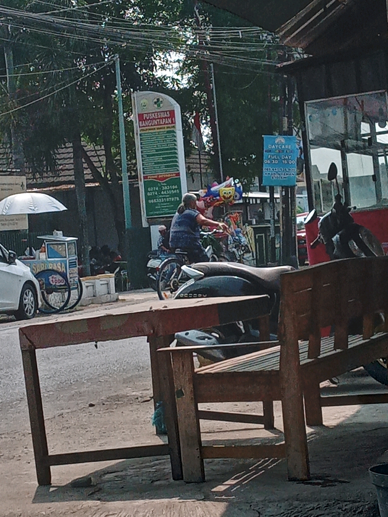 In Banguntapan, Indonesia, a street scene unfolds with a prominent PUSKESMAS BANGUNTAPAN 1 sign, a public health center. To its right, a DAYCARE sign indicates services are available. In the foreground, a wooden bench and table create a seating area. A white car is parked to the left, near a food cart labeled "SARARASA 1000". A man rides a motorcycle in the mid-ground, while other figures are present near a building. Balloons are visible in the upper left portion of the image. The environment suggests a sunny day with some shade from trees and overhead structures.