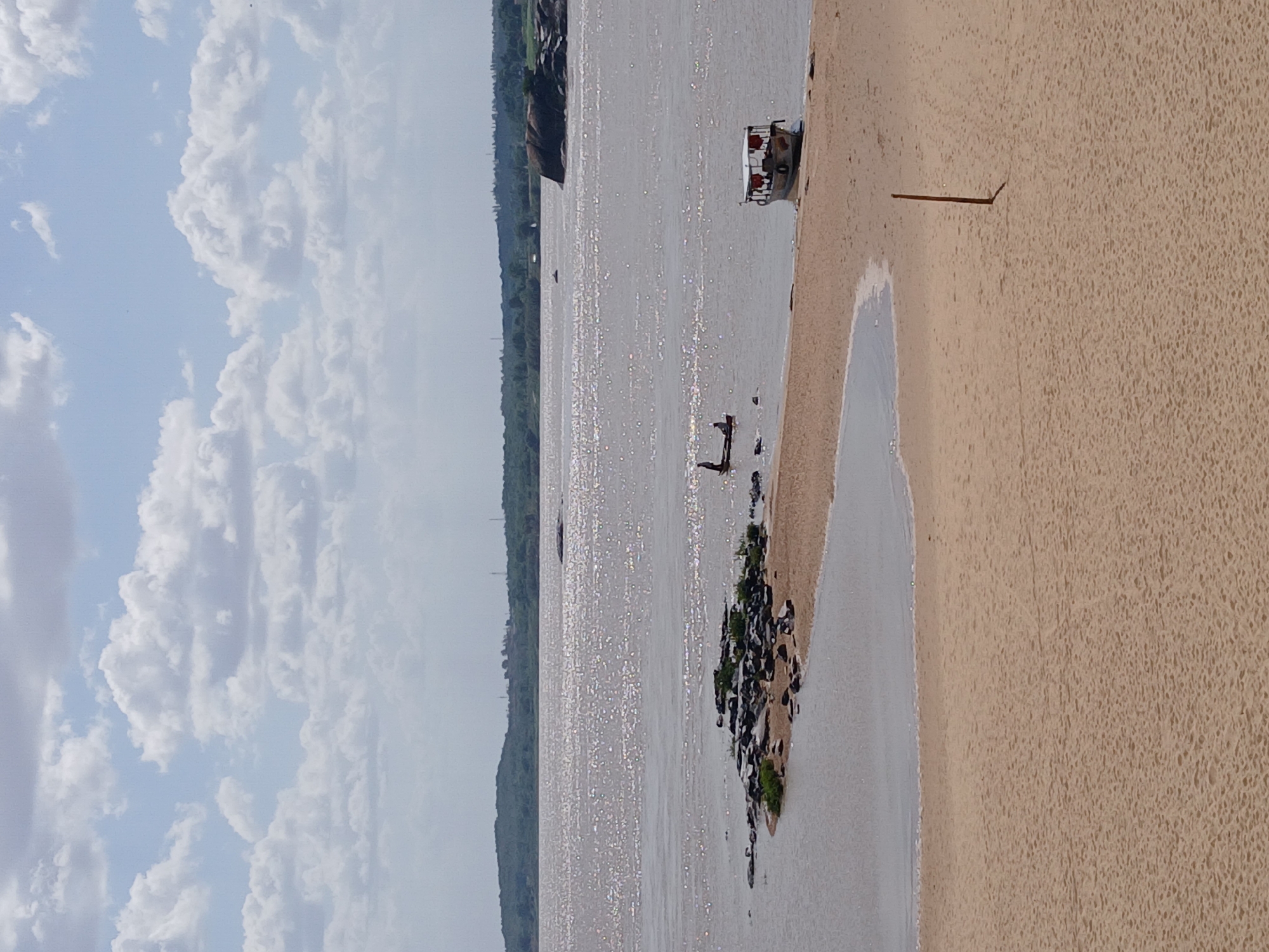 The image depicts a sandy beach next to a body of water, possibly a river or lake. In the distance, there's a stretch of land covered in vegetation. The sky is partly cloudy, with fluffy white clouds dominating the view. A small boat is anchored near the shore, and a lone figure stands in the water. Closer to the beach, there is a cluster of rocks covered in some greenery. A dark line stretches on the sandy ground and seems to be a stick. Based on the request for the location, it seems the image depicts a beach or waterfront in the city of Ciudad Bolivar, Venezuela. However, the specific location within the city cannot be ascertained from the image alone.