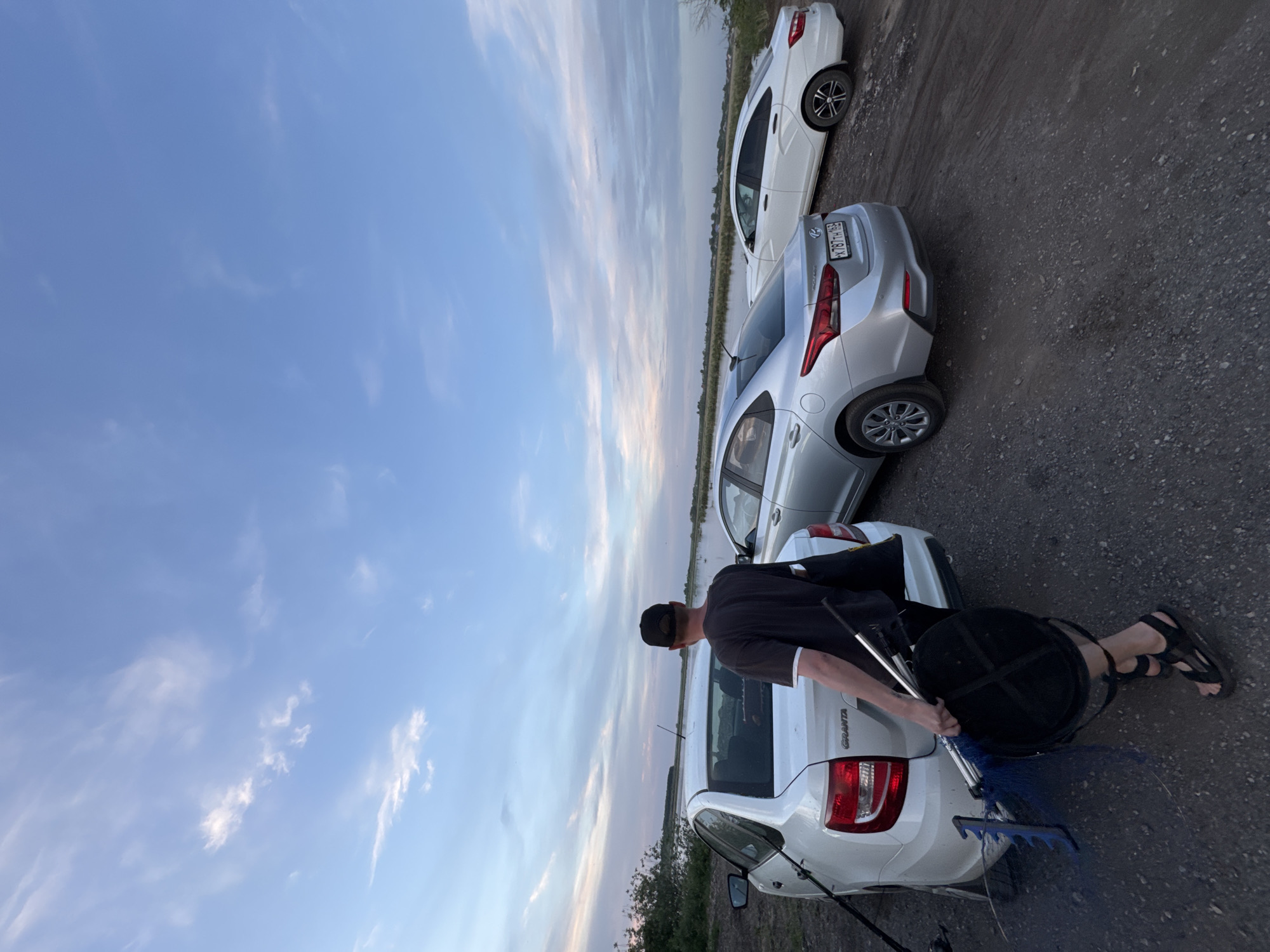 The image shows a landscape view taken outdoors, with a focus on a man and several cars parked along what appears to be a roadside. The sky is visible, with a mix of blue and streaks of clouds, suggesting either early morning or late evening light.
The man is wearing a dark shirt, a baseball cap, and sandals. He is standing near a white car, holding a dark-colored bucket and a fishing net. Another silver car is parked next to the white one, and a second white car can be seen further along.
The area looks like a gravel or dirt roadside next to a body of water, although the water is only subtly visible in the background between the cars. There are some green plants along the road, which suggests a natural environment or the outskirts of a city.
Location information is not present in the image.