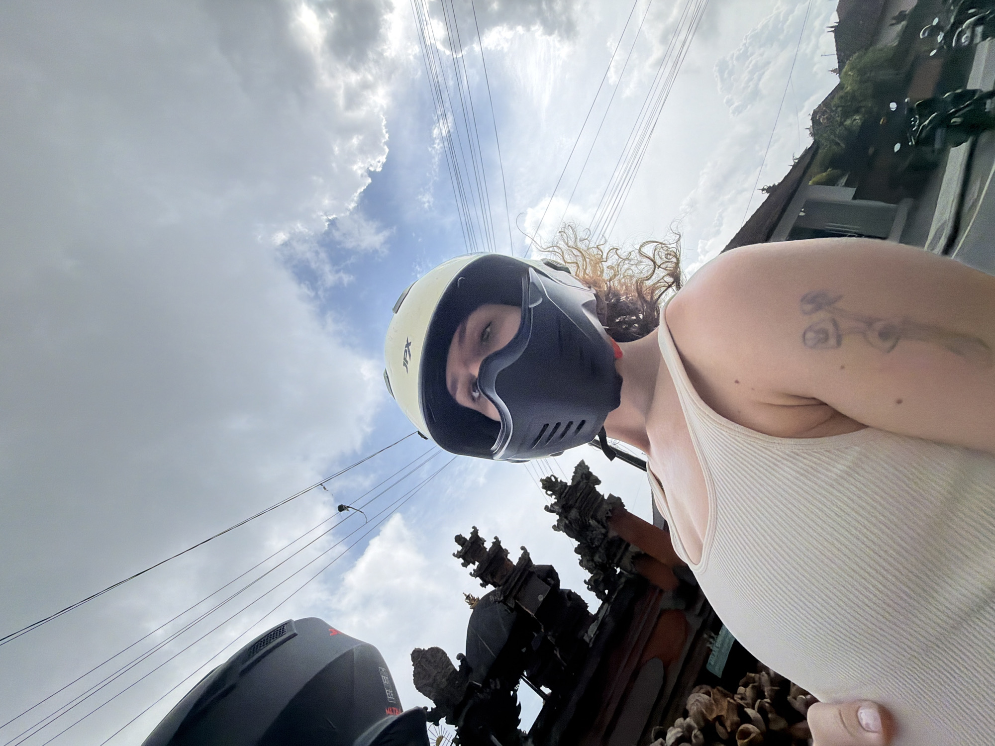 A low-angle, outdoor, waist-up, selfie shot features a person wearing a beige ribbed tank top and a black face mask with a beige motorcycle helmet. The person's right arm is extended towards the camera. The background displays a cloudy sky with power lines, a black motorcycle helmet, and decorative Balinese architecture with carved details, suggesting a location in Mengwi, Indonesia. Multiple parked motorcycles are visible in the distance.