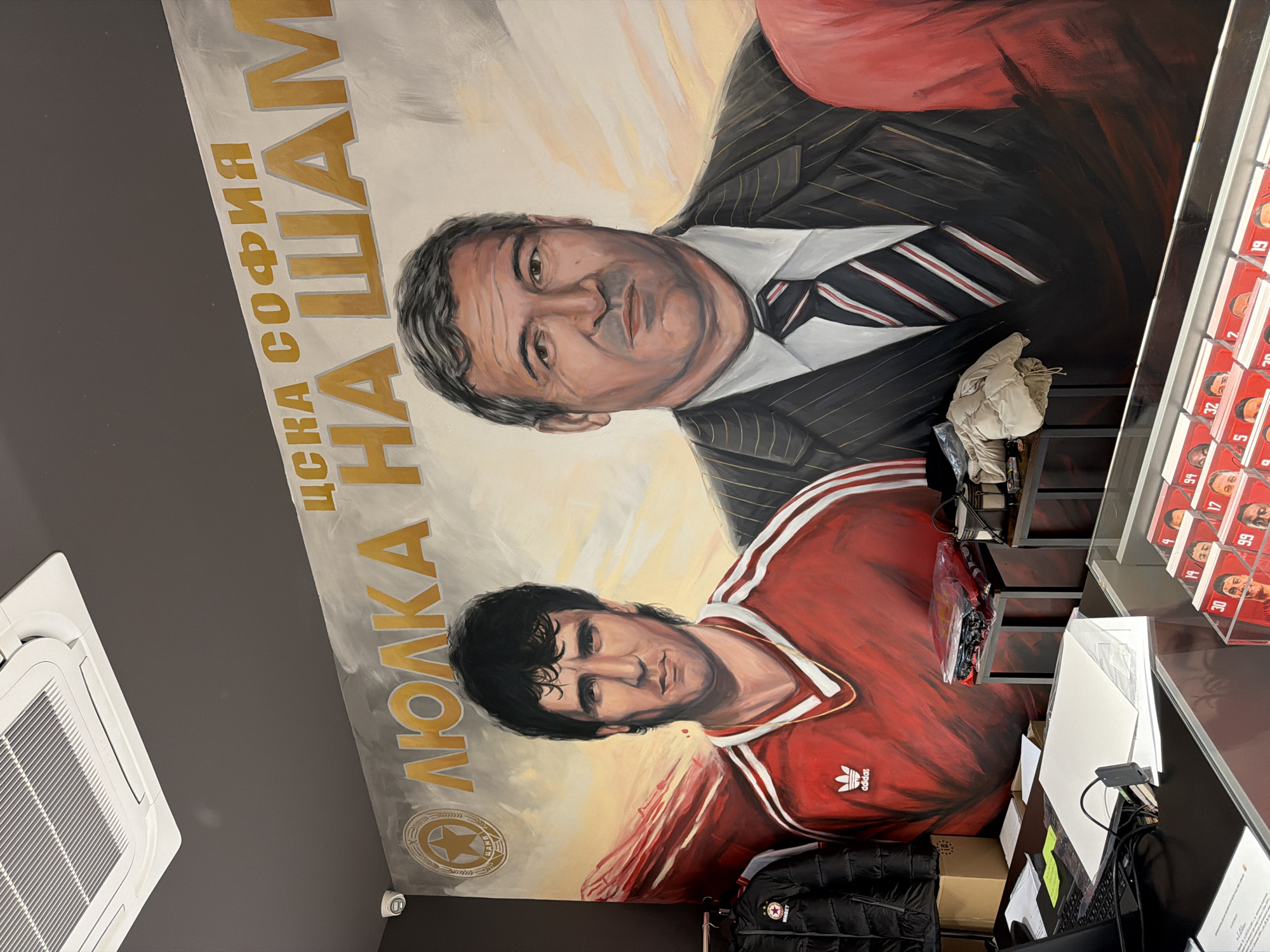 The image, rotated 90 degrees clockwise, displays a large mural on a wall within an indoor setting, likely a fan shop or office related to a sports club in Sofia, Bulgaria. The mural features two prominent male figures. The upper figure is an older man with dark hair, wearing a pinstripe suit and a striped tie. Below him is a younger man with dark, curly hair, dressed in a red Adidas sports jersey with white stripes. The mural's background shows a stylized sky with warm, light colors.
Visible text on the mural includes "ЦСКА СОФИЯ" (CSKA Sofia) and "ЛЮЛКА НА ШАМ" (Cradle of Champions), along with the "adidas" logo on the jersey. These texts strongly identify the mural as a tribute to the CSKA Sofia football club.
To the right of the mural, a counter or desk is visible, equipped with office supplies such as a computer monitor, keyboard, and various papers. A transparent display case on the counter holds numerous small cards or pictures, each featuring a person's face and a number (e.g., "99", "32", "14", "17", "5", "30"), suggesting collectible player cards or merchandise. A light-colored puffer jacket hangs over the counter, and another black jacket with a red star emblem and "WINBET" text is partially visible. On the dark gray ceiling to the left, an air conditioning unit is installed. A small security camera is also present on the wall near the bottom left of the mural.
