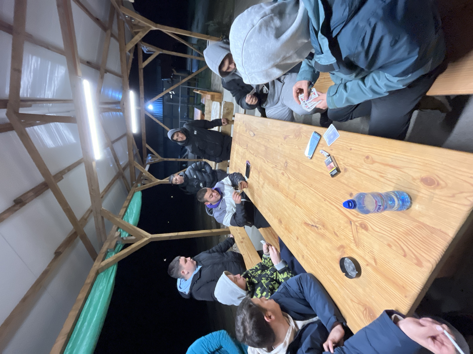 In Karavelovo, Bulgaria, a group of approximately eight young men are gathered around a long, wooden table under a wooden-framed structure illuminated by two fluorescent tube lights. The structure has a partial roof and green tarp siding. The men appear to be engaged in casual conversation, with some holding and playing cards. A mobile phone, playing cards, cigarette pack, water bottle, and an ashtray are on the table. The setting appears to be outdoors at night. The men are dressed in a variety of casual winter clothing, including jackets and hoodies.