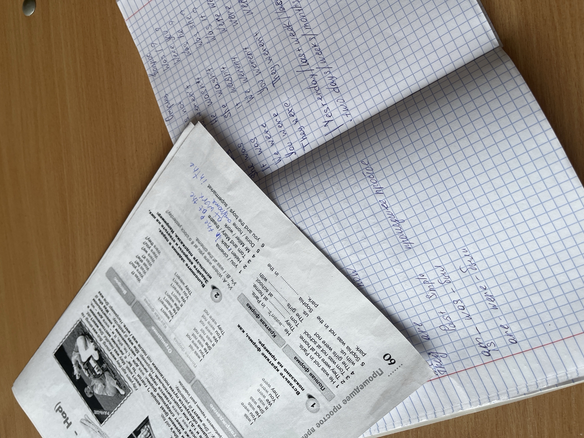 The image shows a desk with English grammar exercises and handwritten notes on notebooks. A worksheet titled "Прошедшее простое время" (Past Simple Tense) is on the left, presenting rules and examples of the verb "to be" in the past tense in Russian. It includes exercises focused on forming positive, negative, and interrogative sentences. A notebook with blue grid paper sits open on the right, containing handwritten notes in English and Russian related to past tense forms of "to be." The notebook and worksheet are resting on a wooden desk.