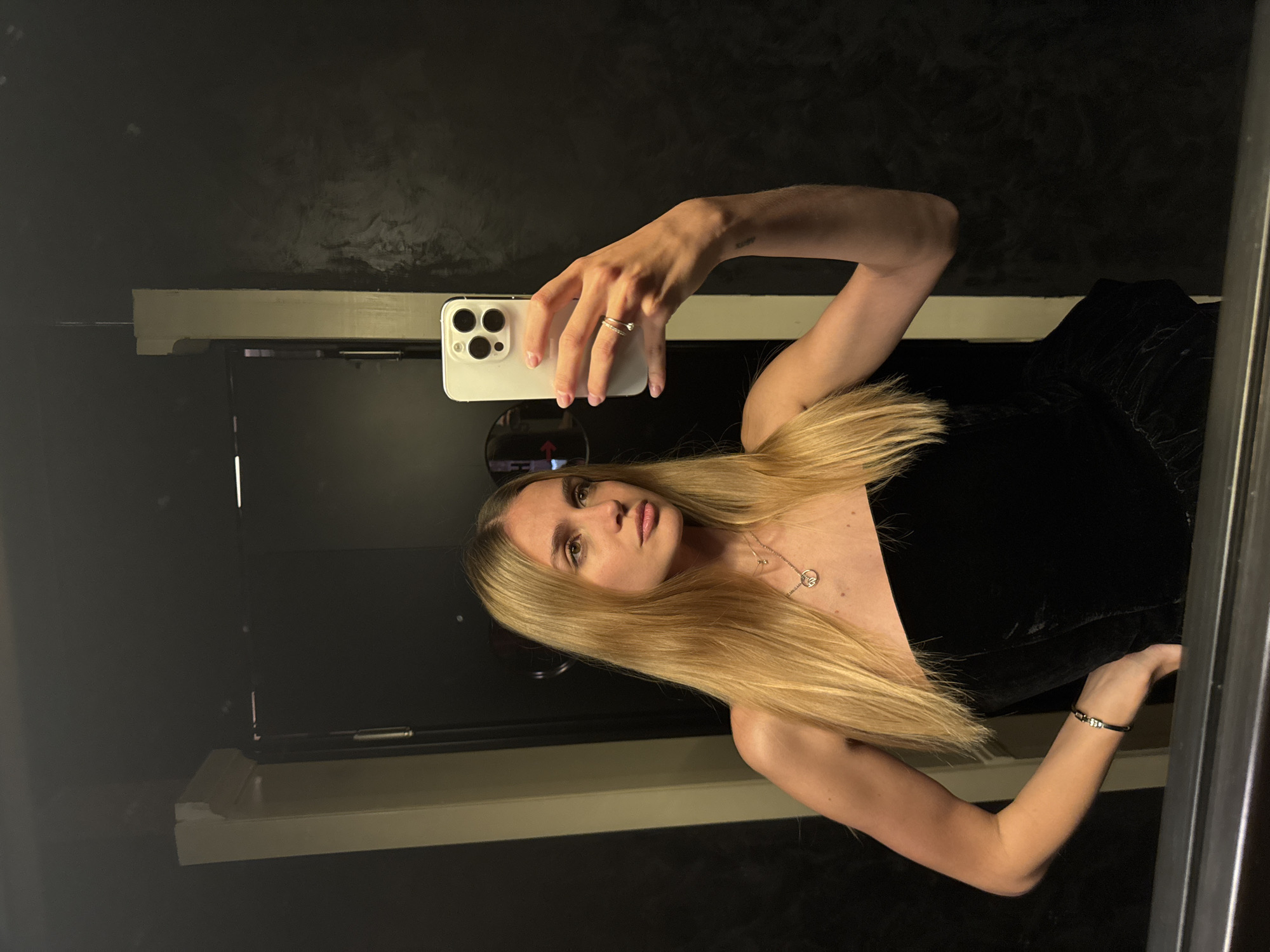 A young woman with long blonde hair takes a selfie in a mirror. She wears a black strapless top and a silver bracelet. She is holding a white iPhone. The background is a dark wall with a light-colored shelf. No information to suggest a location in Tbilisi, Georgia, is discernible from the image.