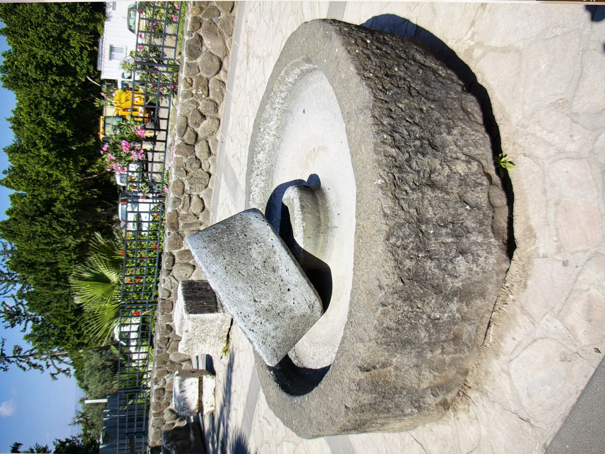 The image shows an ancient stone olive press with a large grinding stone placed on top of a basin. The stone press is located outdoors on a paved ground surrounded by a stone wall. In the background, there is a metal fence, trees, and some vehicles. The scene appears to be part of an archaeological site or museum. This image is not from Kyiv, Ukraine, as the architecture and vegetation do not match that region.