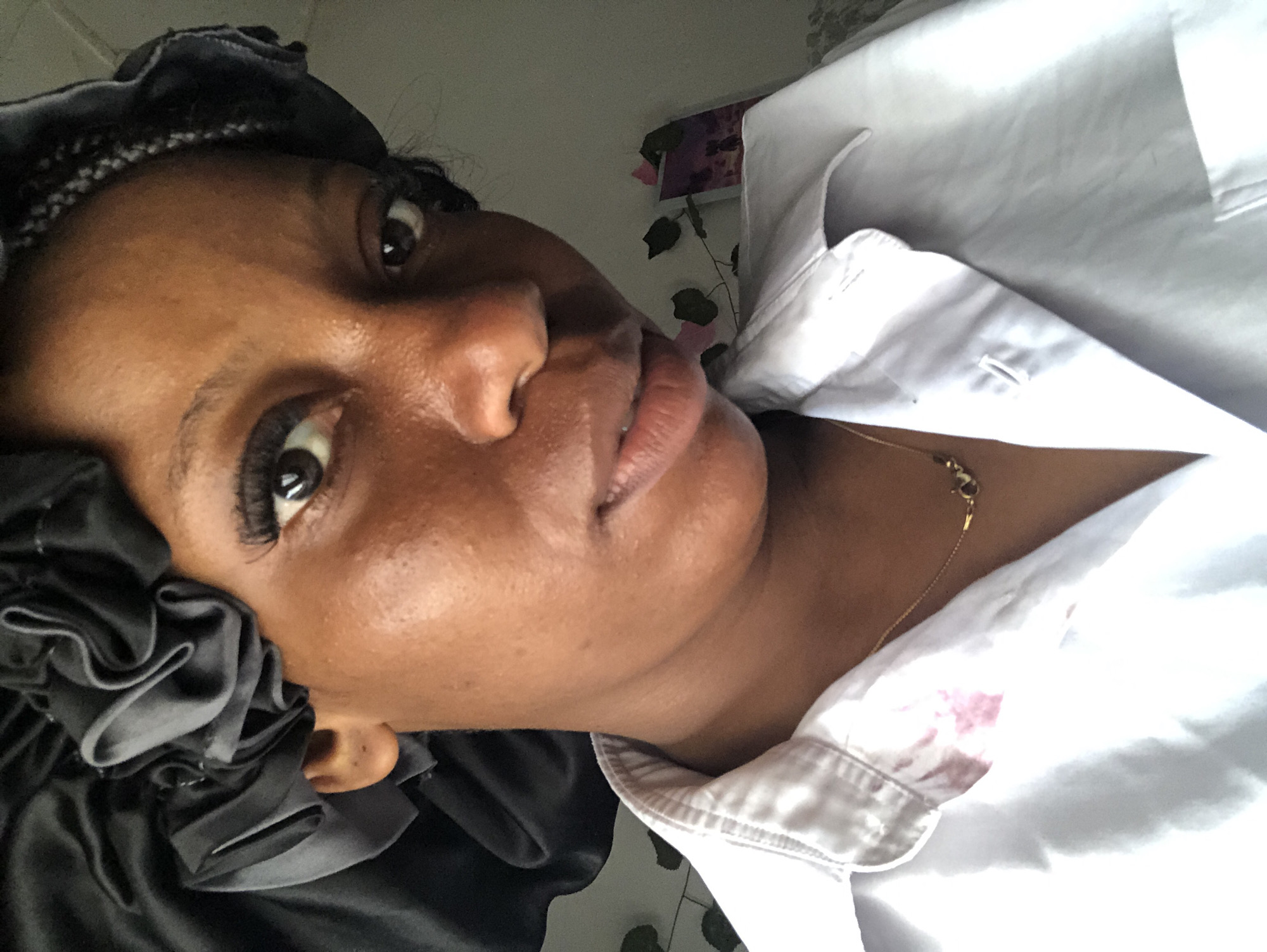 This is a close-up, vertically oriented, selfie-style image of a Black person, likely a woman, with dark skin. She is wearing a black satin bonnet with braided accents around the forehead and a white collared shirt. The shirt has a small, faint pink stain on the right side of the collar. A thin gold chain necklace is visible around her neck. Her eyes are open and looking slightly upwards, and her lips are slightly parted. The image appears to be taken indoors, with a plain wall visible in the background, possibly with some decorative elements such as artificial plants. The lighting suggests it could be daytime, but without further context, the exact time of day is indeterminate. The overall tone of the image is casual and intimate, as is typical of a selfie. There is no visible text in the image.
