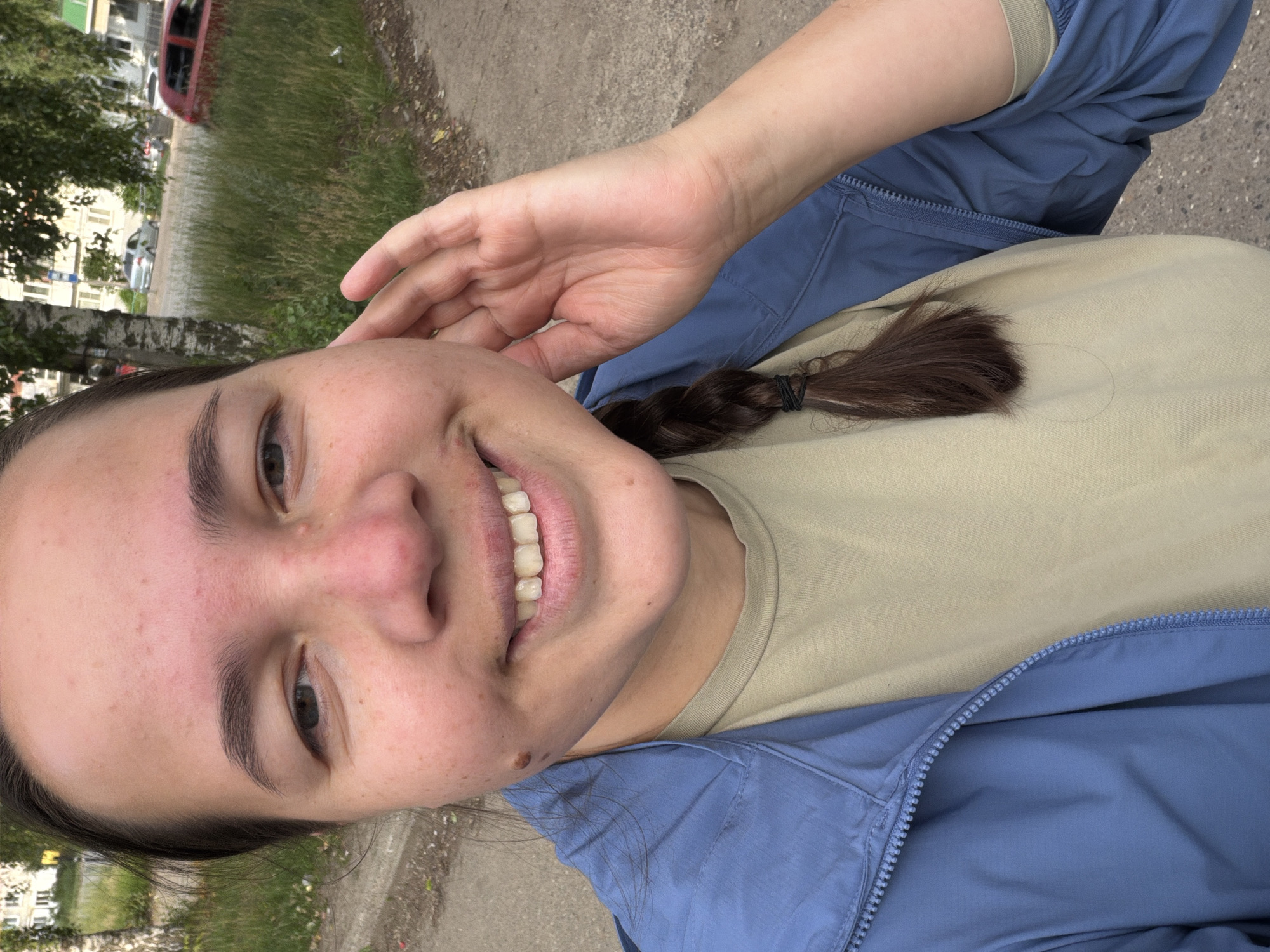 The image shows a close-up, selfie-style shot of a young woman with dark hair, styled in a braid, and fair skin. She is wearing a blue jacket over a beige t-shirt. Her hand is raised to her ear. The background shows a street scene with a grassy verge, a sidewalk, and a few vehicles, including a red car. The presence of birch trees and the architectural style of the buildings visible in the background suggest an urban or semi-urban environment in Russia. Specifically, the distinct style of the buildings and the overall appearance of the street are consistent with locations in the city of Chaikovsky, Russia. The lighting indicates it is daytime, likely overcast.