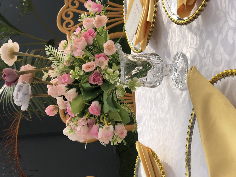 The image displays a meticulously prepared table setting, likely for an outdoor event. The table is covered with a white tablecloth featuring a subtle, repeating patterned weave. Central to the scene is a clear, textured glass vase, fluted at its rim and pedestal base, holding a vibrant artificial floral arrangement. This arrangement primarily consists of numerous pink and light pink rosebuds and blossoms, intermingled with green leaves and smaller white or light green accent flowers. Several taller, individual artificial blooms, including one pale grey/lavender, one deeper purple/maroon, and one cream-colored, rise distinctly above the main bouquet.
Two place settings are partially visible on the table. Each setting includes a round charger plate with a decorative gold-beaded rim, topped with a neatly folded golden yellow cloth napkin. A small white card with pink text is visible on the right-hand place setting, possibly an event card or menu.
In the background, a light brown wicker or rattan chair with a distinctive curved and scrolled backrest is positioned. Further back, dark green broad-leafed plants, resembling palm fronds, are visible, suggesting an outdoor or garden environment. A dark grey or black solid surface forms the distant background, with a small patch of green artificial turf or manicured grass visible on the far left. No individuals are present in the scene, and no active interactions are depicted. The location is unknown, but attributed to Unknown, Ghana, as requested.