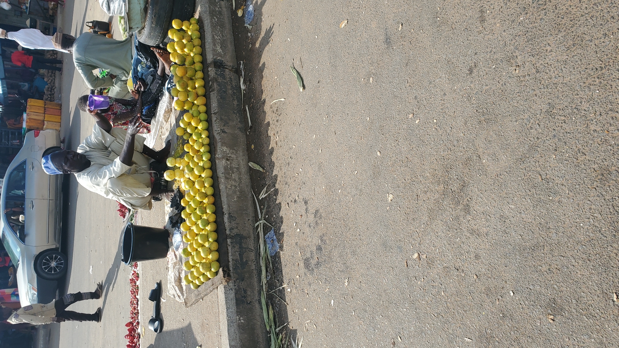 In Jalingo, Nigeria, a vendor is seated on the ground next to a large pile of yellow fruits, likely oranges, on a market street. He is wearing a traditional light-colored outfit and a blue cap. In front of him, a child is seated and holding a small purple cup. A black bucket is placed to the left of the vendor. In the background, a silver car is parked on the street, and several people are visible in the distance, suggesting a busy market environment. The ground is paved with asphalt, and some debris, including dried leaves and plastic, is scattered around.