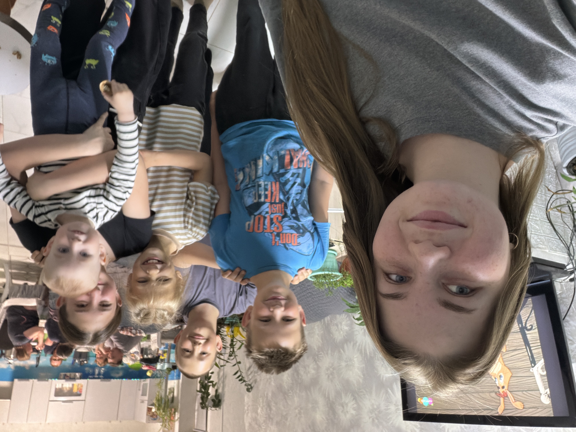 The image depicts a group of five children and a young woman lying on their backs in what appears to be a modern kitchen. The woman is positioned at the right edge of the frame, with her face visible. The children are lined up from left to right, their heads near the bottom of the image. The children are wearing various casual clothes. The kitchen features white cabinets, countertops, and a tile floor. A TV displaying a cartoon-like image is visible in the background, near the woman. There are plants visible in the background of the image. The exact location cannot be definitively determined as Большой Букор from this single image.