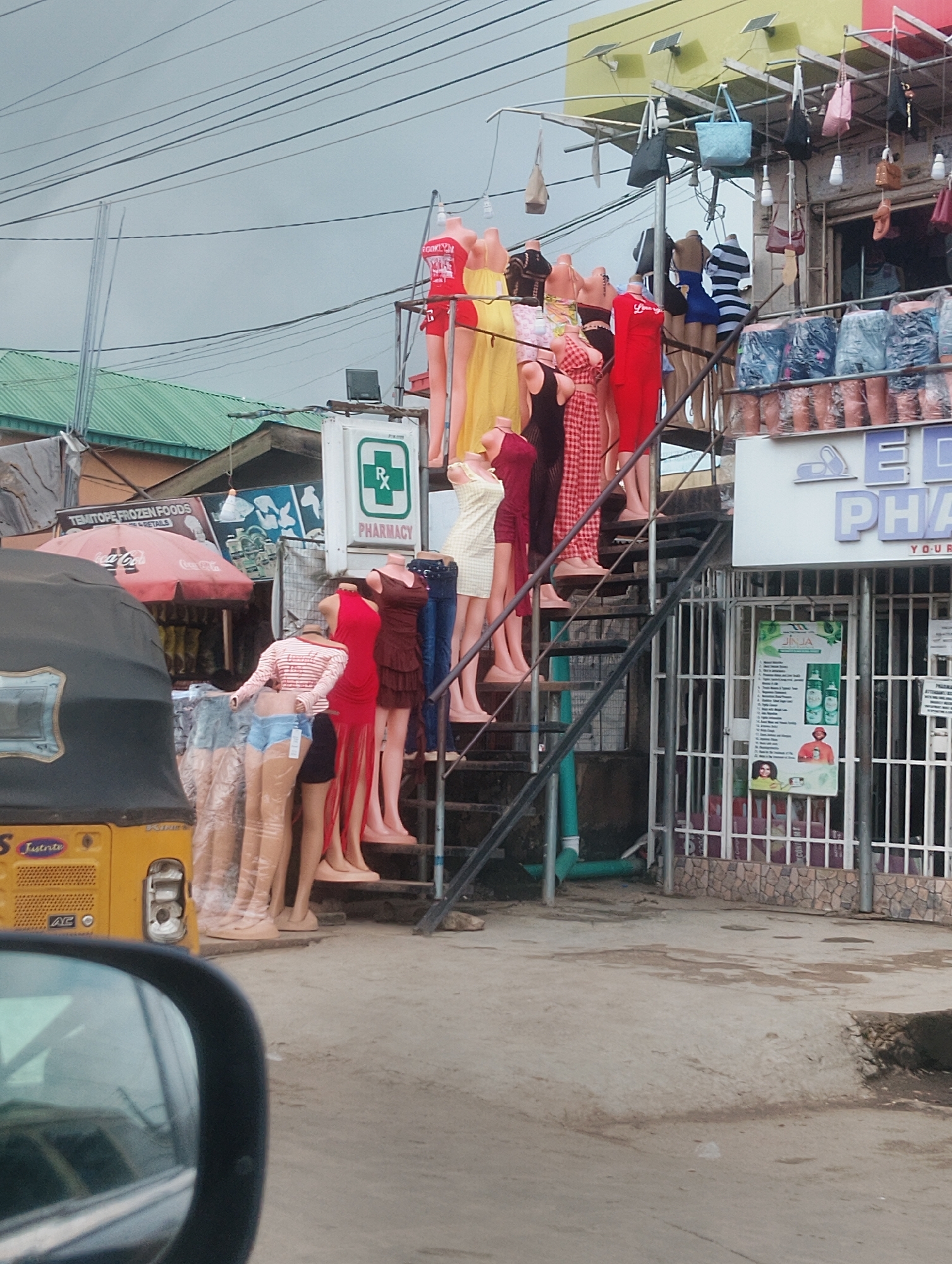 This image shows a street scene in Iju, Nigeria, likely during daytime, with an overcast sky suggesting potentially cloudy weather. The foreground is dominated by a yellow three-wheeled vehicle, possibly a tuk-tuk or motorcycle taxi, partially visible from the driver's perspective, indicated by the reflection in a side mirror.
The main focus of the image is a collection of mannequins displayed on a metal staircase leading up to a shop. These mannequins are dressed in various clothing items, including dresses, shorts, and tops, suggesting a retail setting for fashion. The mannequins are arranged in a densely packed manner, creating a visually striking display.
Behind the mannequins, several shop fronts are visible. One sign clearly reads "PHARMACY" with a green Rx symbol, indicating a drugstore. Another sign above it has the partial text "AED PHA YOUR", likely part of a larger business name or advertisement. Further back, a sign for "TEMITOPE FROZEN FOODS" is also visible, along with a red Coca-Cola umbrella, hinting at a market or commercial area. Bags and clothing are also displayed hanging from an overhead structure. The overall scene depicts a busy street with commercial activity centered around clothing and pharmacy businesses.