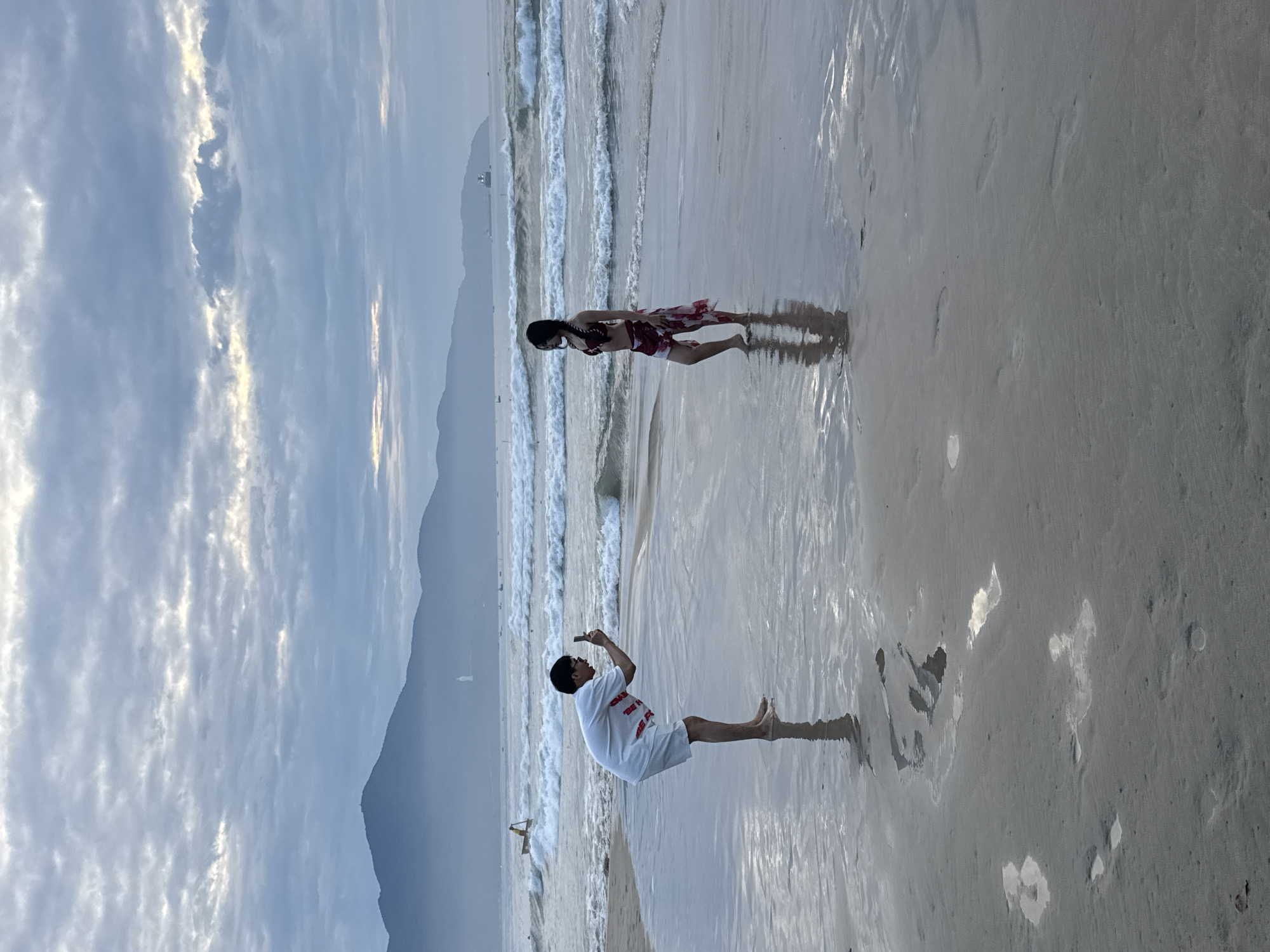 Two people are on a beach in Đà Nẵng, Vietnam. A man is taking a photo of a woman who is standing in the shallow water near the shoreline. The beach is sandy, and the ocean is calm with small waves. A mountain range is visible in the background under a cloudy sky.