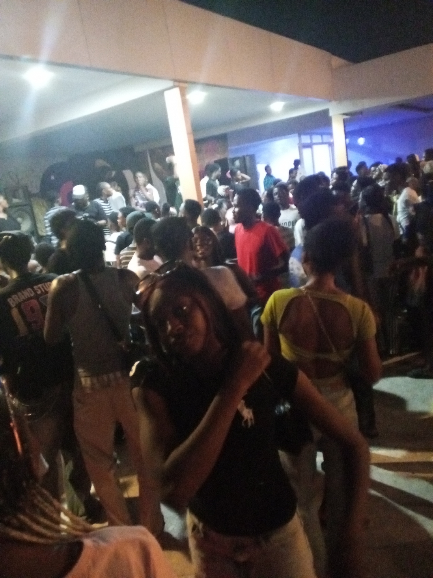 A crowd of people is gathered in what appears to be a public space in Adankolo, Nigeria. A building with visible columns and lighting is in the background. The people are dressed in casual clothing. In the foreground, a woman is facing the camera with her arm raised. The overall setting suggests a social event or gathering.
