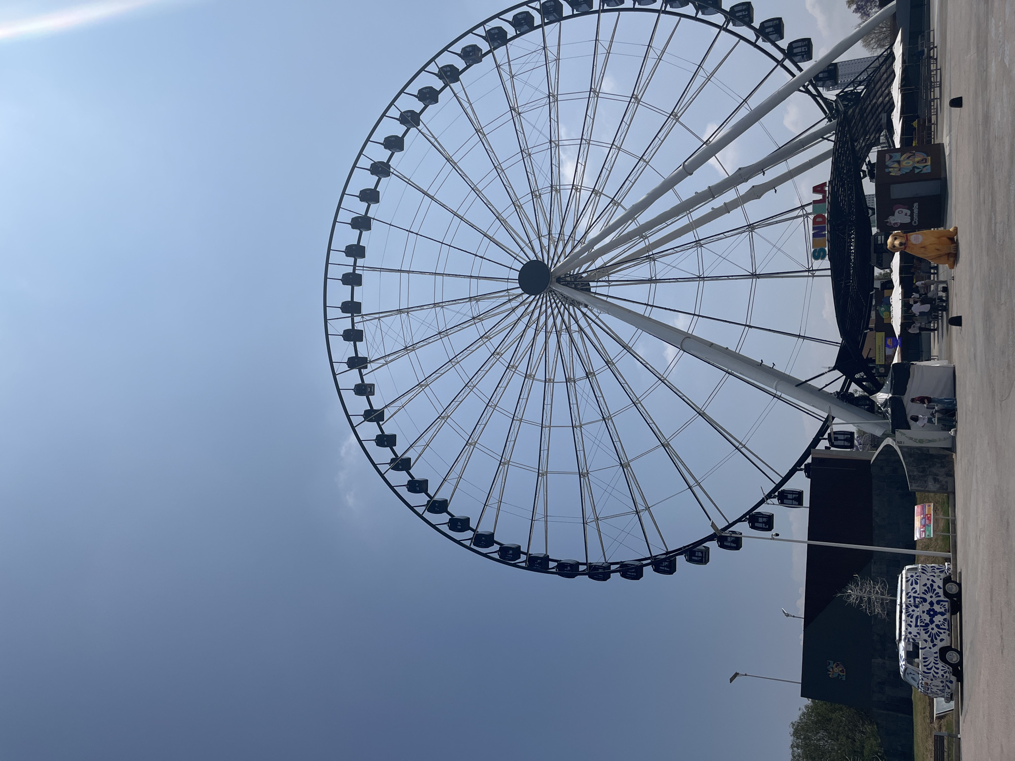 The image depicts a Ferris wheel in Puebla, Mexico, seen from a low angle, with a clear blue sky above. The Ferris wheel has black passenger cars and is supported by white beams. At ground level, to the right of the Ferris wheel, there is a structure with a banner that reads "SINDLA". A person in a lion costume stands nearby. There is also a van with blue and white patterned decoration.