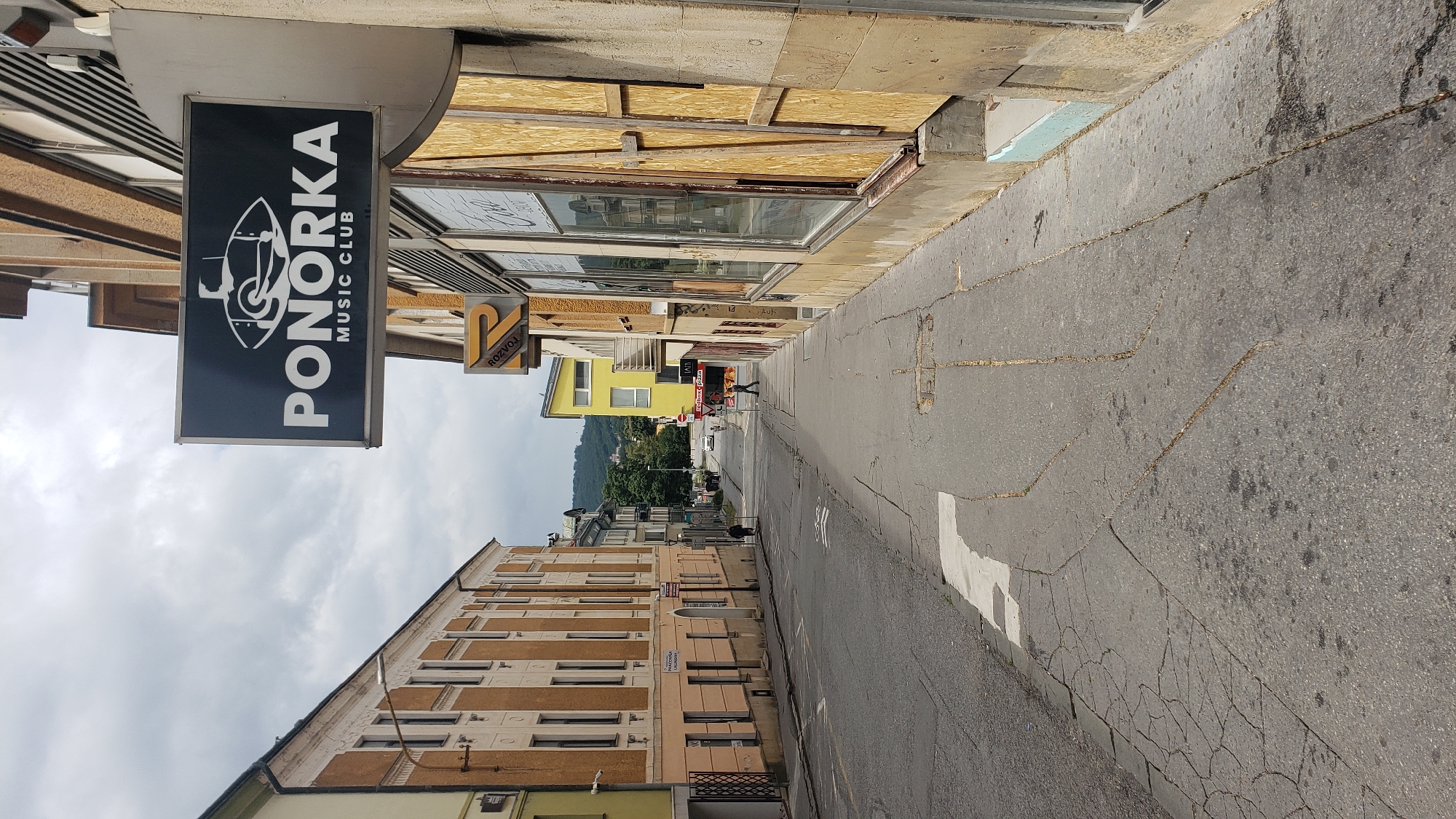 The image depicts a street scene in Prešov, Slovakia. A black sign with white lettering reading "PONORKA MUSIC CLUB" is prominently displayed, accompanied by a stylized eye logo. The sign is attached to a building on the right side of the street. The building appears to have boarded-up windows, suggesting it may be closed or undergoing renovation. To the left of the street, there is a building with a yellow and orange facade, featuring multiple windows. Further down the street, a yellow building stands out, and a distant castle or fortress is visible on a hill. Several cars are parked or driving on the street, and a few pedestrians are walking along the sidewalk. The road surface is asphalt and shows signs of wear and tear, with visible cracks and patches. The sky is overcast with a dramatic cloud formation.