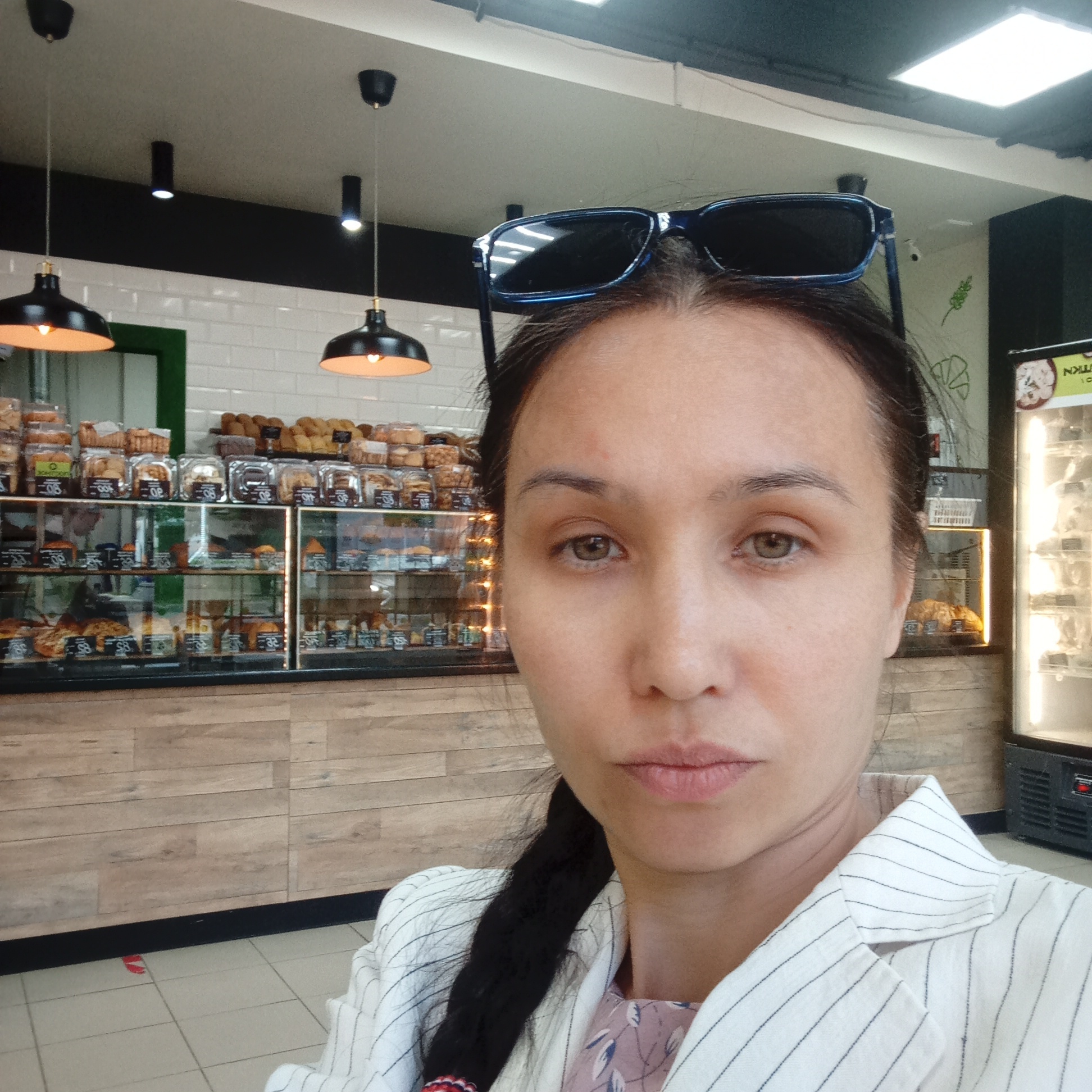 A woman is taking a selfie in what appears to be a bakery or food establishment in Izhevsk, Russia. She is wearing a white jacket with thin black pinstripes and sunglasses perched on her head. The background shows display cases filled with various baked goods, including pastries and bread, with price tags visible. Hanging light fixtures are also present in the scene. The overall environment suggests a retail setting, likely a bakery or cafe.