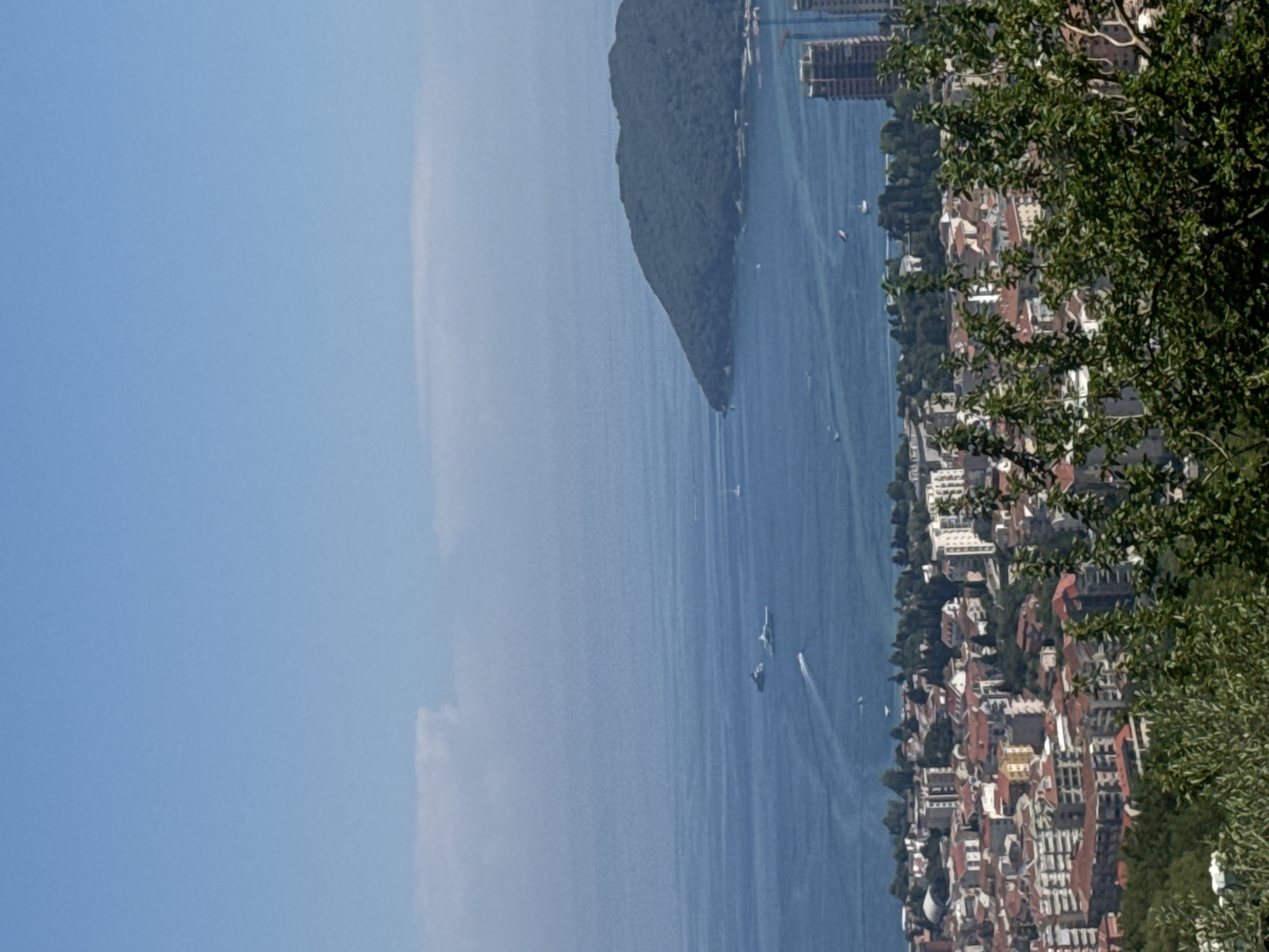 An aerial view captures the coastal city of Podostrog, Montenegro. The foreground features lush green foliage and trees. Beyond the trees, a densely populated urban area with buildings of varying heights and architectural styles, characterized by red-tiled roofs, stretches towards the sea. Several boats are visible on the calm blue water, and a prominent, tree-covered island juts out into the sea on the right side of the frame. The sky is a clear blue with a few wispy clouds.