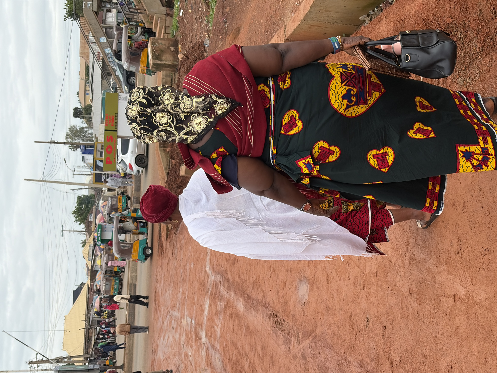 The image depicts an outdoor scene in Gura Top, Nigeria, likely during the daytime given the bright, overcast sky. The ground is a reddish-brown dirt, common in many parts of Nigeria, and a dusty road runs through the scene, lined with small shops and various vehicles.
The primary subjects are two individuals. One is a woman wearing a dark green dress with a colorful pattern featuring yellow and red hearts. She has a red headscarf and a black headwrap adorned with gold floral embroidery. She is carrying a black handbag and a wallet in her right hand and wears a blue wristband. She appears to be walking or standing. Next to her is another person wearing a white garment and a red headscarf, facing away from the camera.
In the background, a busy street scene unfolds. Several auto-rickshaws, known locally as Keke Napep, are visible, some parked and others in motion, indicating a bustling environment. Stalls and shops with awnings and umbrellas line the road, suggesting a market or commercial area. Various people are seen in the background, some walking and others near the vehicles. One shop sign is partially visible, displaying "POS" in large red letters, and a phone number. The overall atmosphere suggests a typical day in a Nigerian town or city, with everyday commerce and transportation in full swing. The weather appears to be overcast or cloudy, as there are no strong shadows, and the sky is a uniform grey.