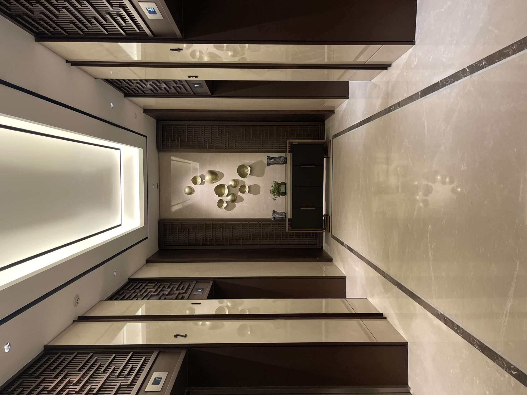 This media file depicts an interior scene, likely a hallway or lobby within a modern residential or commercial building. The perspective is looking upwards, creating a sense of height and grandeur. The dominant materials are polished light-colored marble for the flooring and walls, contrasted with dark wood paneling and ornate, lattice-like wooden screens.
The scene features several architectural elements. A long, illuminated rectangular light fixture is prominent on the ceiling. Along the walls, dark wood cabinets and decorative screens are integrated. In the central area, a dark cabinet with gold accents stands against a patterned wooden wall, adorned with a cluster of gold, bowl-shaped decorative objects. A small potted plant adds a touch of greenery.
There are no people visible in the image. The activity, if any, is implied to be the passage through this space. The lighting suggests it is daytime, or the interior is well-lit, creating a bright and luxurious ambiance. The location context is South Jakarta, Indonesia, which could imply a tropical climate influencing the architectural design for ventilation and aesthetics, though this is not directly evident from the image itself.
Notable details include the geometric patterns of the wooden screens and the clean lines of the overall design. The marble flooring features subtle veining, and a dark, thin marble strip runs along the floor at an angle, delineating a pathway. On the upper levels of the hallway, recessed panels with buttons are visible, likely for elevator controls, suggesting this might be near an elevator bank. One panel has a blue button with a symbol that might indicate a "door close" or "up" function, and another has a symbol possibly related to a service call. Another panel has a number "1" clearly visible. The overall impression is one of sophisticated and contemporary design.
