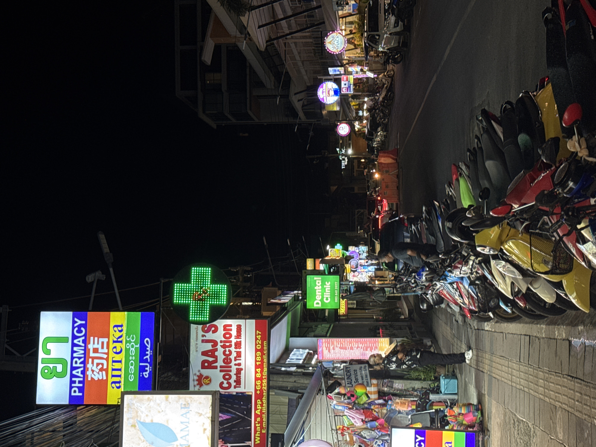 An eye-level, full shot captures a street scene at night in Karon, Thailand. The left side of the frame features a row of brightly lit signs, including a tall, multi-colored sign for a pharmacy with text in Thai, English, Chinese, Russian, Burmese, and Arabic. Below this, a sign reads "MAME" with a blue leaf graphic. Further down, a sign for "RAJ's Collection" advertises tailoring and Thai silk. Adjacent to the pharmacy sign is a green, illuminated cross, a common symbol for pharmacies. To the right of the signs is a sidewalk with a man in a black floral shirt and black pants standing with his arms crossed, facing away from the camera and towards the street. On the right side of the frame, the street is lined with parked motorcycles of various colors. Buildings with multiple stories and illuminated shopfronts are visible along the street. Vehicles, including cars and more motorcycles, are present on the street, suggesting ongoing activity. The overall atmosphere is that of a lively commercial street at night.