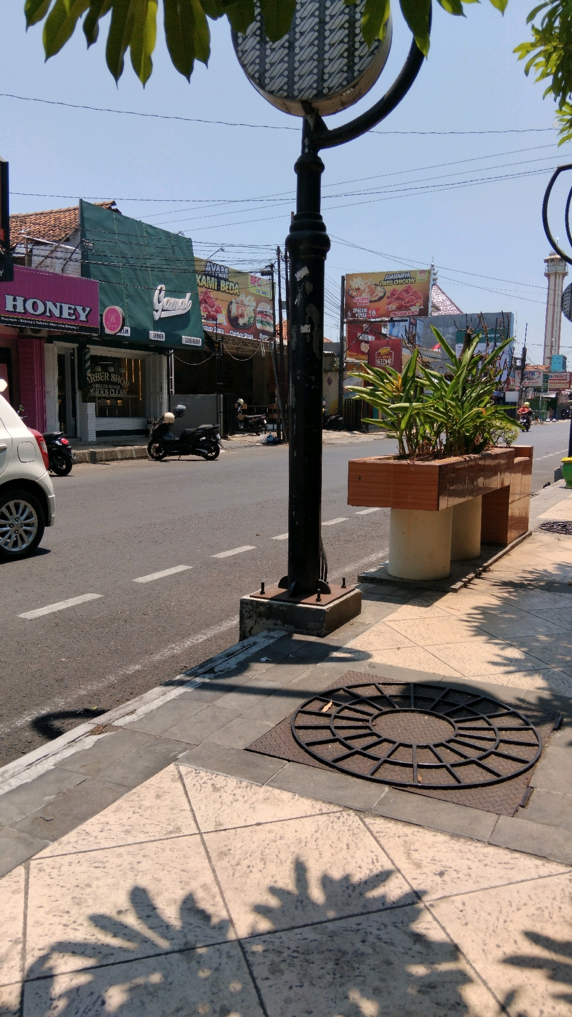 The image depicts a street scene in Batang, Indonesia. The foreground shows a sidewalk with patterned tiles and a large circular grate set into the ground, casting shadows from overhead foliage. A dark lamppost stands prominently on the sidewalk, bolted to a concrete base. To the right, a raised planter box with a wooden exterior and concrete cylindrical supports holds lush green plants. The street itself is paved and has white lane markings. On the street, two motorcycles are visible; one parked on the side and another in motion. A white car is parked on the left side of the street. Businesses with various signs, including "HONEY" and a barber shop, line the street. Billboards advertising food items like fried chicken are also present. In the distance, a minaret of a mosque can be seen. The sky is clear and blue, indicating a sunny day.