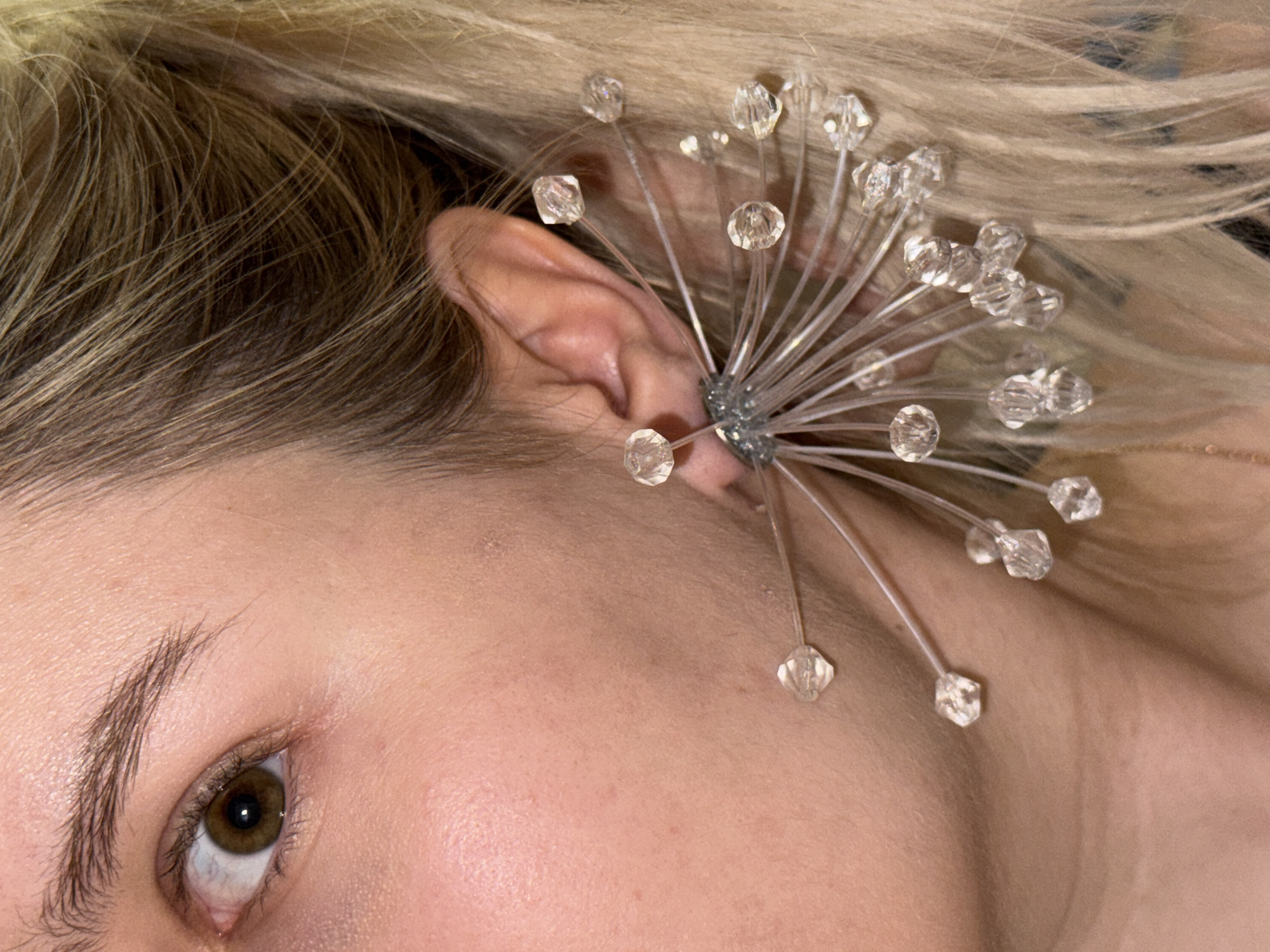 The image is a close-up, eye-level shot of a person's face and ear, focusing on a unique, spiky earring adorned with clear, faceted beads. The earring is silver and starburst-shaped, with thin wires radiating outwards, each tipped with a bead. The person has light-colored skin, a brown eye with a surprised expression, and blonde hair. The background is blurry and appears to be indoors, with some hints of dark clothing. The specific location within a city is unknown.