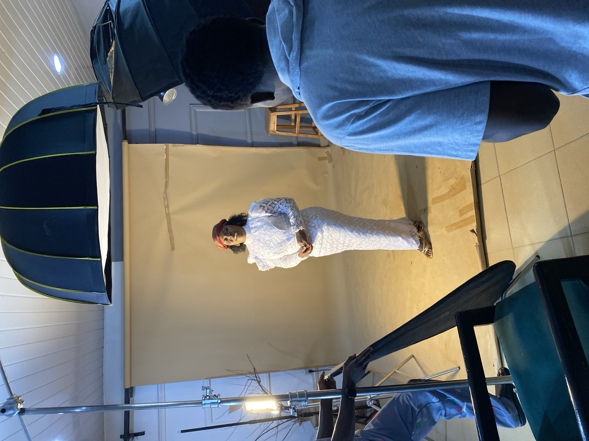 An indoor photography studio setup is depicted, captured from an elevated perspective looking down into the scene. The image is rotated 90 degrees clockwise, showing a vertical composition.
In the center, a woman stands facing the camera (implied to be the viewer's position). She is wearing a long, elegant white lace dress with long sleeves and a red headscarf. Her dark hair is visible, and she has a gentle expression, appearing to smile slightly. Her hands are clasped in front of her. She is positioned against a seamless light beige/tan paper backdrop that extends from the wall to the floor, creating a continuous surface.
To the upper left, a person, likely the photographer, is visible from behind, wearing a blue hooded sweatshirt. They are positioned to capture the woman's photo. Another individual is partially visible in the bottom right corner, wearing a patterned garment, seemingly adjusting or operating equipment.
The studio is equipped with professional lighting. A large, dark blue softbox is prominent on the left, providing diffused light. Another softbox is partially visible at the top right. Light stands support the equipment, and an exposed light bulb or strobe is visible on a stand at the bottom left. A wooden stool is in the background behind the woman, and a chair with a blue seat and dark frame is in the foreground on the bottom right. The floor is tiled, and the ceiling appears to have white paneling with circular light fixtures.
The scene captures a portrait photography session in progress. No discernible text is visible in the image. The setting suggests a professional studio environment located in Bukuru, Nigeria.