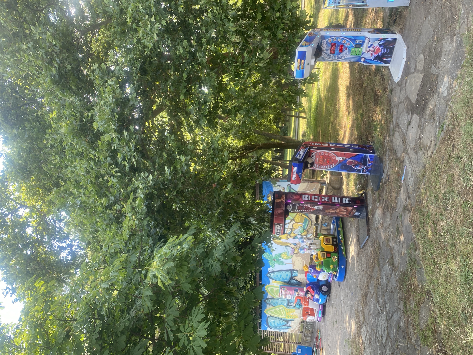The image shows a children's amusement area in a park in Burgas, Bulgaria. Several cartoonish amusement rides for young children, including a train and cars, are lined up in front of a colorful graffiti-covered wall. To the right of the rides, there are three arcade-style boxing machines. The ground is paved with irregular stones and patchy grass. Lush green trees with dense foliage form the background, with sunlight filtering through the branches, suggesting a bright, sunny day. The overall scene depicts a playful, outdoor entertainment space within a natural setting.