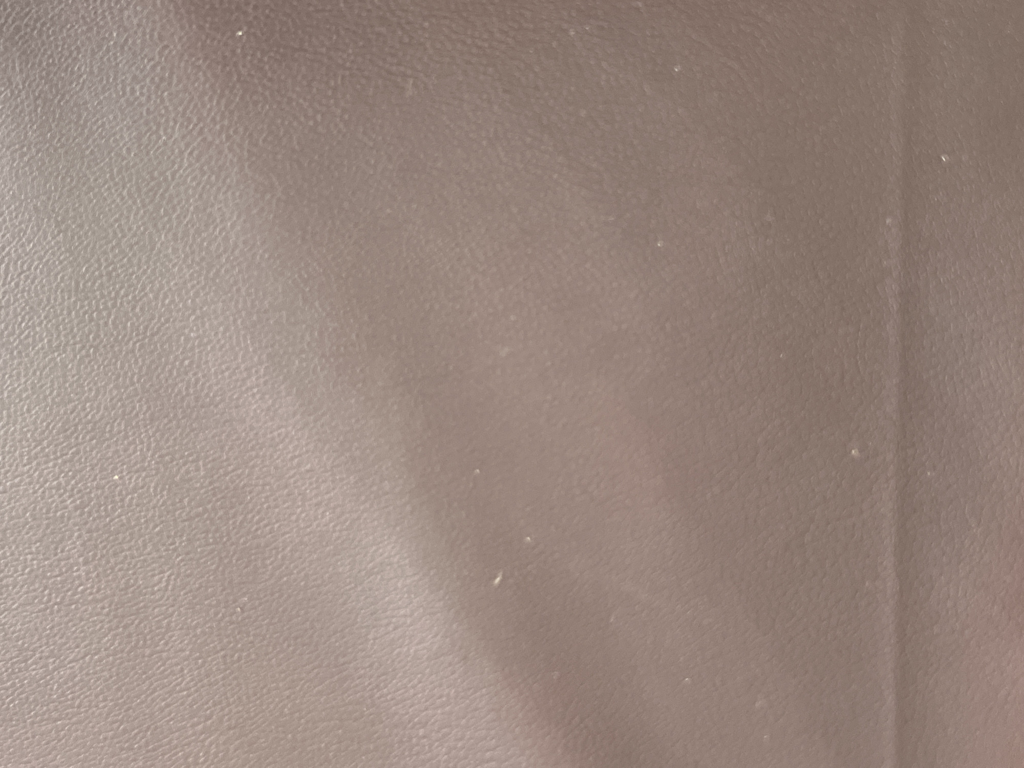 The image presents a close-up, forensic view of a textured surface, characterized by a muted purplish-brown or taupe coloration. The material exhibits an irregular, pebbled grain pattern, consistent with aged leather or a synthetic equivalent. Subtle variations in ambient lighting create gentle topographic undulations across the surface, indicating a non-planar form. Numerous small, light-colored particulate specks, consistent with dust or textile fibers, are sparsely distributed across the texture. No individuals, specific objects beyond the surface material, actions, or interactions are discernible within the frame. The location is Casablanca, Morocco.