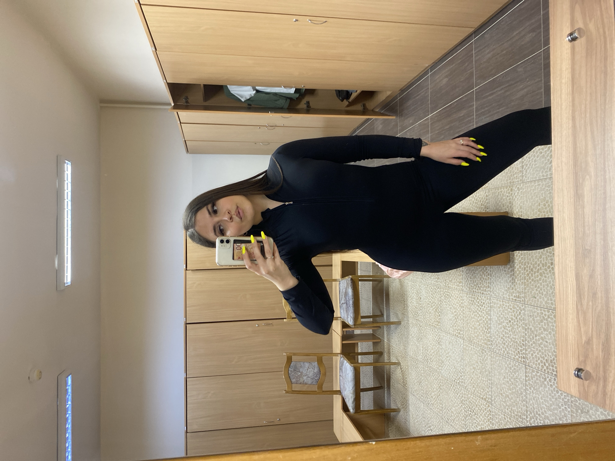 A young woman is depicted taking a mirror selfie in what appears to be an indoor changing room or locker facility. She stands with her body slightly angled, facing the reflection of her smartphone. She has long, straight, dark brown hair and is dressed in a form-fitting, long-sleeved black jumpsuit. Her left hand, featuring bright neon yellow nail polish and a ring on her ring finger, holds a white smartphone with a small graphic or sticker displaying "00" on its case. Her expression is neutral, with her lips slightly parted.
The background, visible in the mirror's reflection, consists of light-colored wooden lockers or cabinets lining the walls, some of which are partially open. Two wooden chairs with upholstered seats are present near the bottom of the frame. The floor is covered with small, light-colored speckled tiles. A rectangular artificial light fixture is visible on the ceiling, illuminating the plain, light-colored walls. The setting suggests a functional space like a gym, school, or workplace changing area located in Chaikovsky, Russia. The time of day is not indicated, as the scene is lit by artificial light. No other discernible text is visible in the image.