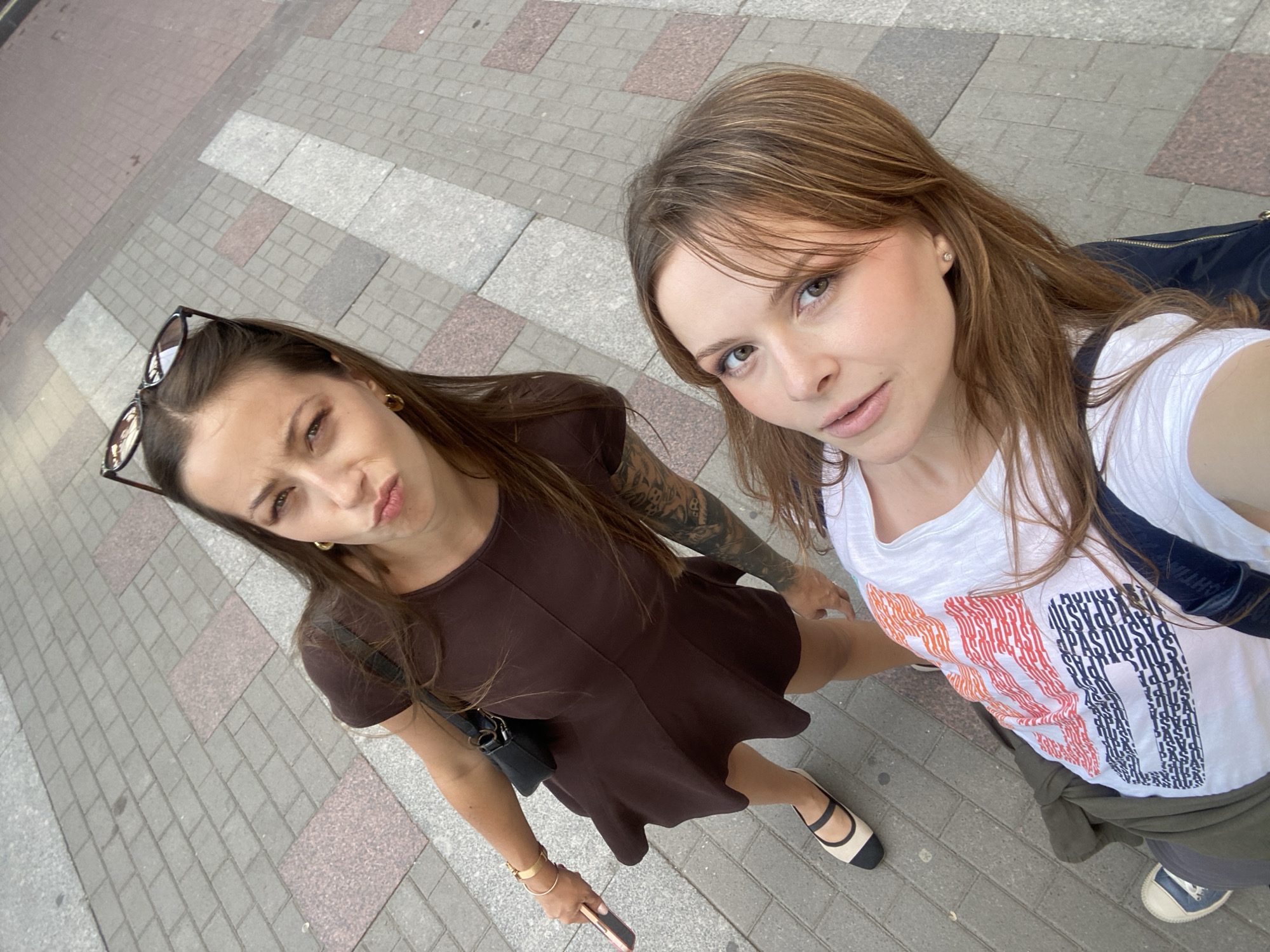 This image depicts two women posing for a high-angle selfie photograph on a paved pedestrian surface in Sinyavino, Russia, during daylight hours.
The woman on the left has long brown hair, with dark sunglasses resting on top of her head. She wears a dark brown, short-sleeved, flared dress, and gold hoop earrings. Her left arm displays multiple gold bangles, and she holds a light-colored smartphone in her left hand. A prominent dark tattoo sleeve is visible on her right arm. She is wearing cream-colored ballet flats with black toe caps and straps. Her expression features puckered lips.
The woman on the right, who appears to be holding the camera, has long, light brown hair with bangs. She wears a white t-shirt adorned with a red and blue graphic pattern. A dark green jacket is tied around her waist, and a dark bag strap is visible over her right shoulder. A dark-colored sneaker with a white toe cap is partially visible on her left foot. She has a small stud earring in her left ear and looks directly at the camera.
The ground beneath them is a paved surface consisting of rectangular light gray and darker gray blocks, interspersed with larger, rectangular reddish-brown granite or stone slabs arranged in a linear pattern.
