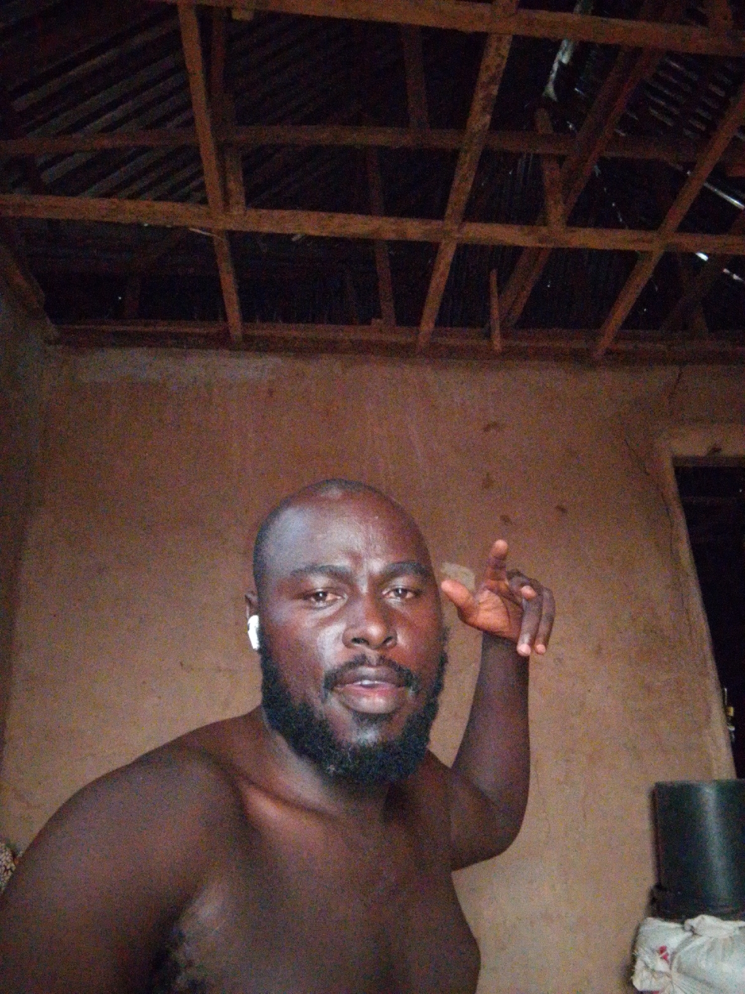 A man with dark skin, a shaved head, and a black beard is in the foreground. He is shirtless and has a white wireless earbud in his left ear. He is partially raising his right hand. The background consists of a textured brown wall and a wooden frame ceiling with corrugated metal sheeting visible through the gaps. A partially open doorway is visible to the right, behind the man, with a black object near it. The location is identified as Mulke Barikin Sayejo, Nigeria.