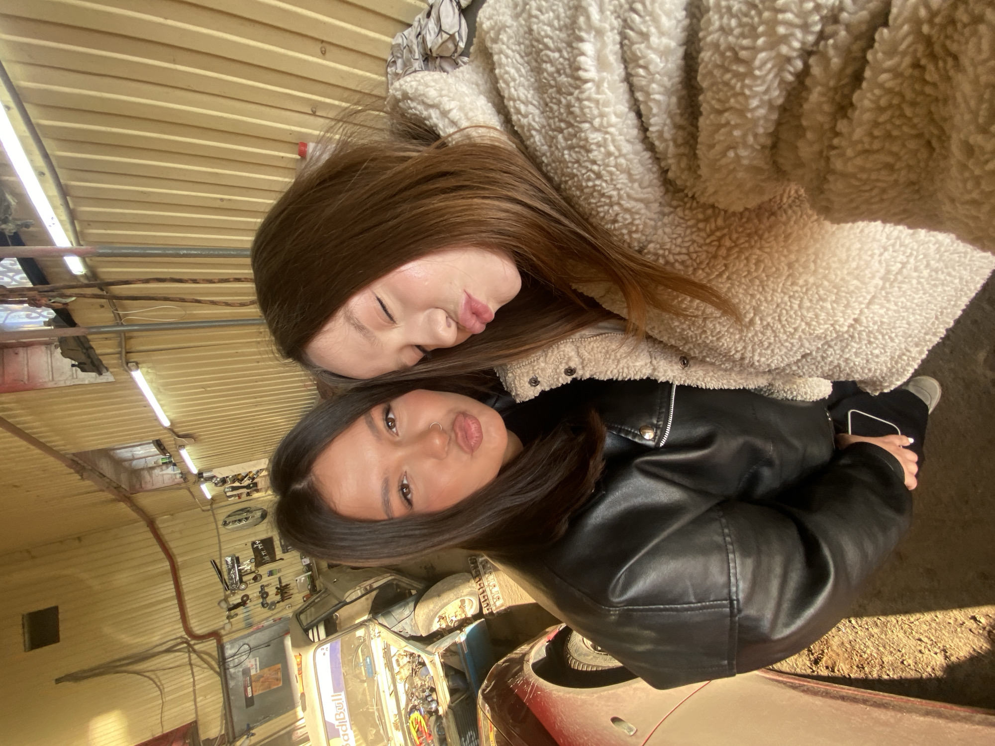 Two young women pose closely together in an auto repair garage. The woman on top is wearing a cream-colored fuzzy jacket and has long brown hair. She is making a kissing face. The woman below is wearing a black leather jacket and also has long brown hair. She is also making a kissing face. In the background, there are several cars in various states of repair, tools hanging on the wall, and fluorescent lights. The garage floor appears to be made of concrete. The specific location is Chaikovsky, Russia.