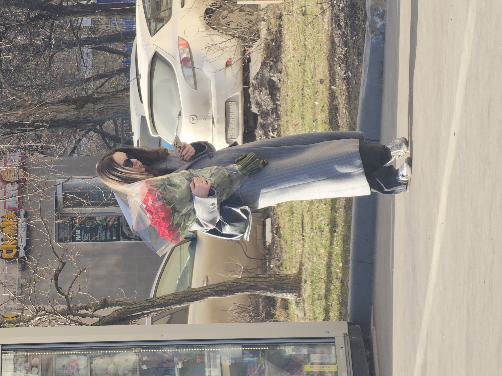 An outdoor scene in Khimki, Moscow Oblast, Russia, during daylight hours under clear weather. A single female subject stands slightly off-center, facing forward, with her head tilted down, looking at a smartphone held in her right hand. She has long dark hair, wears sunglasses, a long grey pinstriped coat, dark trousers, and light-colored sneakers. A black shoulder bag hangs from her left shoulder. She holds a large bouquet of red roses, encased in clear plastic wrapping, against her left side. To the subject's left is a light-colored kiosk or newsstand with items visible through a glass display, including a magazine with "НАРЛЕИКИ" on its cover. The immediate ground is concrete pavement with a blue-painted curb. Behind the subject, a strip of green grass and bare-branched trees are visible. Parked vehicles include a white Mazda 3 hatchback, seen from the rear, with a visible license plate "О 676 РН 150," and a partially obscured gold-colored sedan. In the background, a grey building displays signage in Cyrillic, including "РЫБА" (Fish) and "СКЛАД" (Warehouse/Store).
