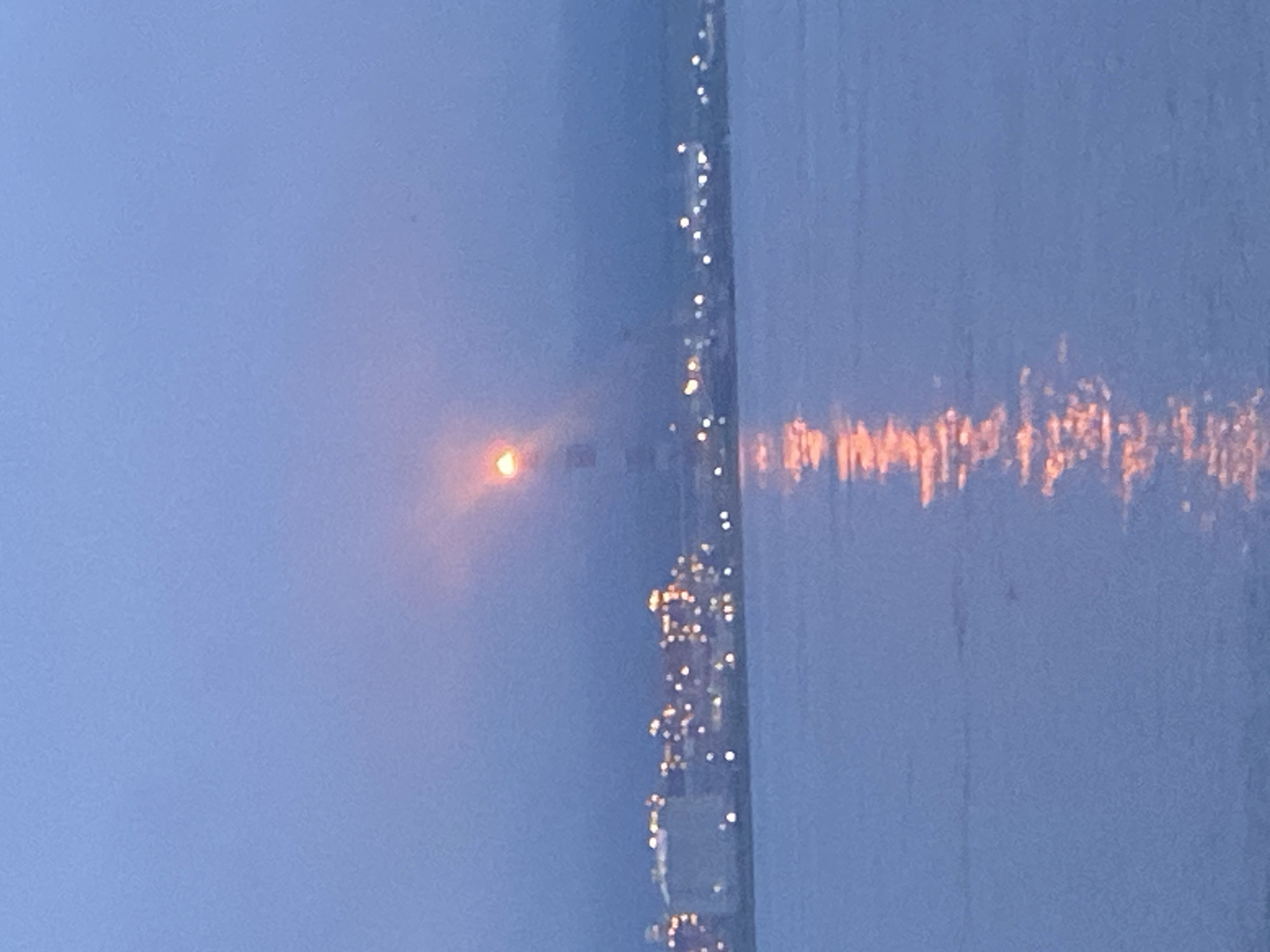 The image captures a twilight scene with a lighthouse at its center, illuminated by what appears to be the moon or a bright light at its apex. The lighthouse stands on a shoreline with a city skyline in the background, characterized by a cluster of buildings with numerous lights, suggesting an urban environment. The water in the foreground reflects the ambient light, creating shimmering patterns on its surface. The overall atmosphere is serene and slightly hazy, typical of dawn or dusk. The location is identified as Korsakov, Russia. There are no visible people or animals in the frame.