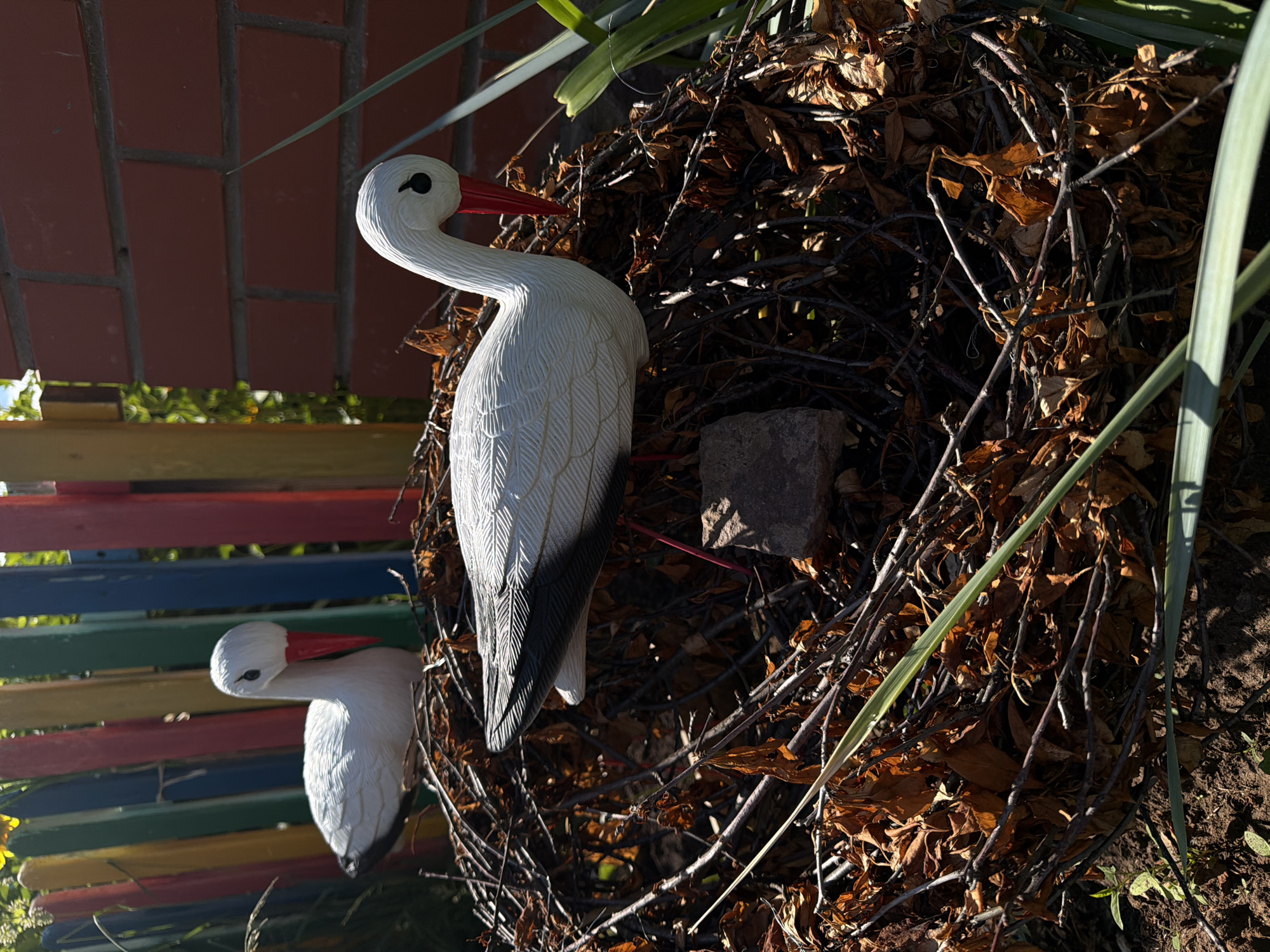 Two white stork figurines are positioned in a nest made of twigs and dried leaves. The nest rests on the ground in an outdoor setting. Behind the nest is a colorful wooden fence with vertical slats painted in red, yellow, green, and blue. To the right of the nest, a brick wall is visible. The scene is illuminated by sunlight, casting shadows and highlighting the textures of the figurines, nest, and surrounding elements. The overall impression is a decorative garden display.