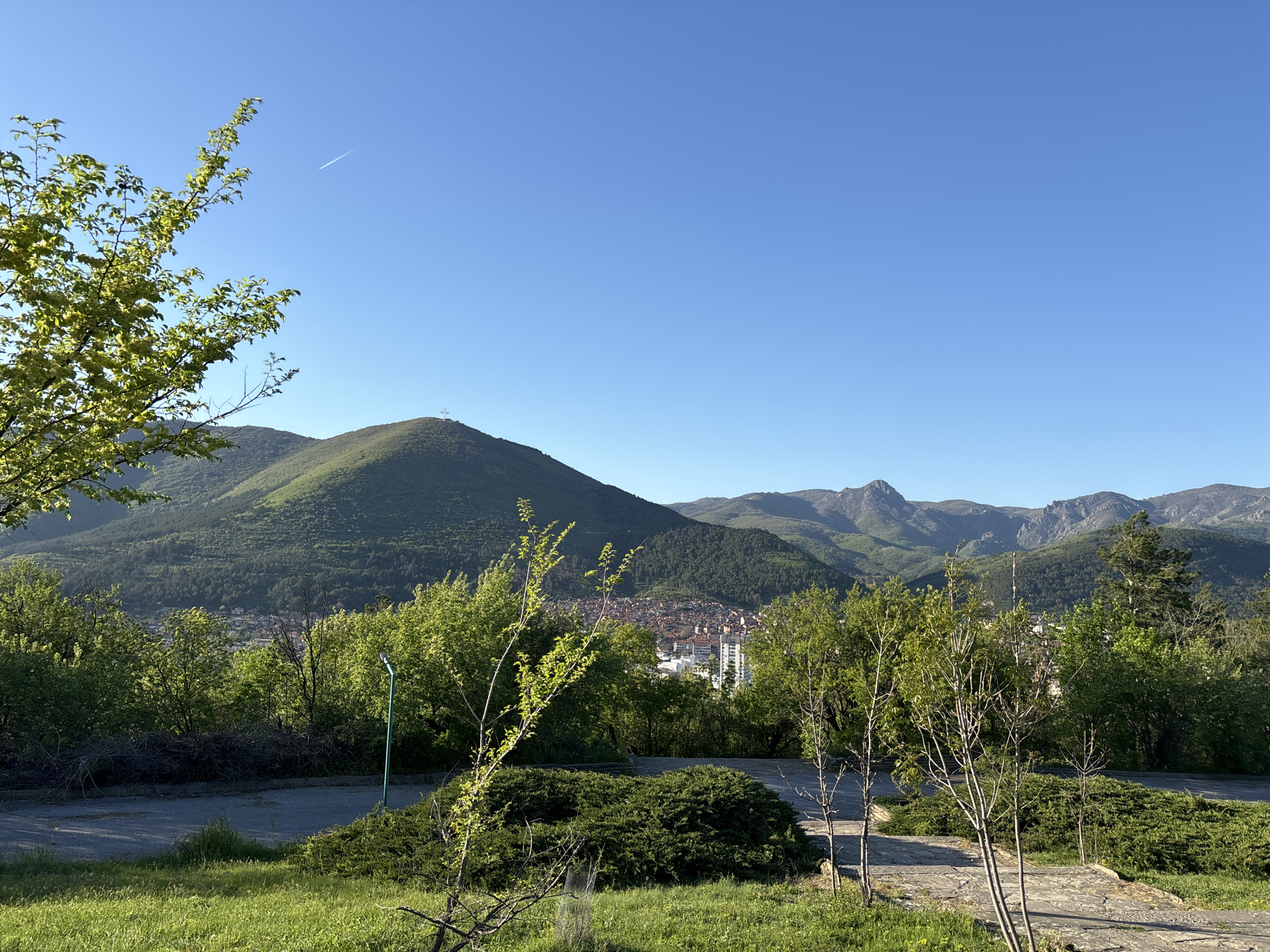 The image depicts a landscape scene in Plovdiv, Bulgaria. In the foreground, there is a grassy area with young trees and bushes. A paved pathway is visible in the mid-ground. The background features a prominent green hill with a cross at its peak, likely the Alyosha Monument on Bunarjik Hill. In the distance are more mountains, partially obscured by the trees in the mid-ground, and a portion of the city's buildings is visible between the foliage. The sky is clear and blue. There appears to be a contrail from an airplane high in the sky.