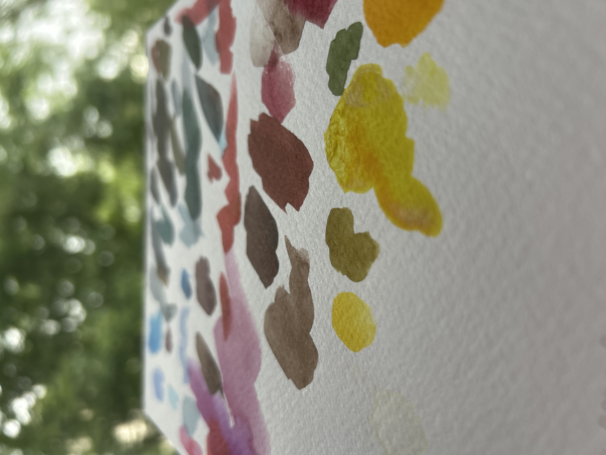 The image shows a close-up of a watercolor painting on textured white paper. The painting consists of dabs of various colors including yellow, green, brown, pink, purple, and blue-gray. The background is blurred and appears to be foliage, possibly from trees. The location in Sofia, Bulgaria is not apparent from this image.